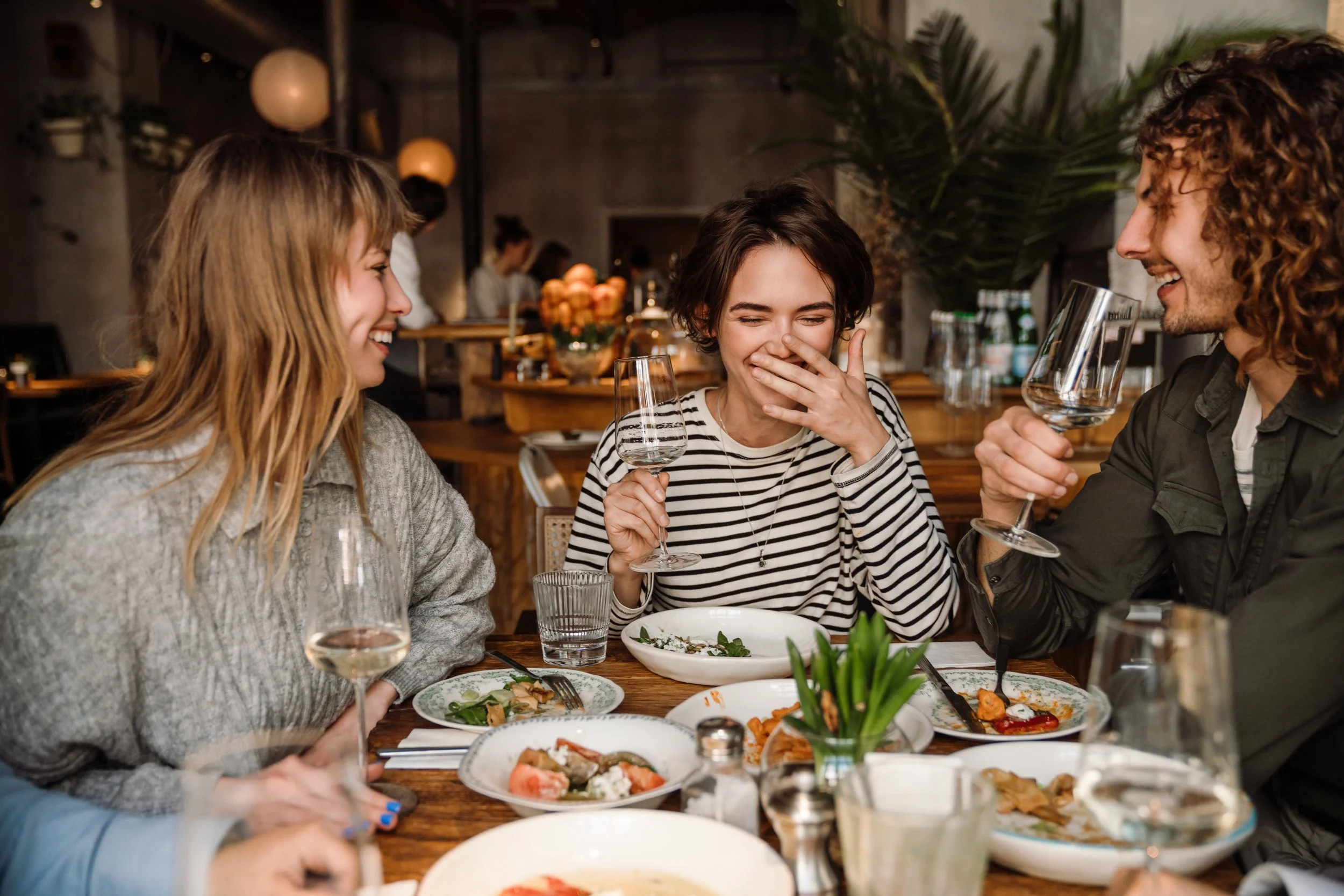 Four friends are sitting at a restaurant table, laughing and enjoying drinks and food. There are various plates of food, glasses of wine, and a salad on the table.