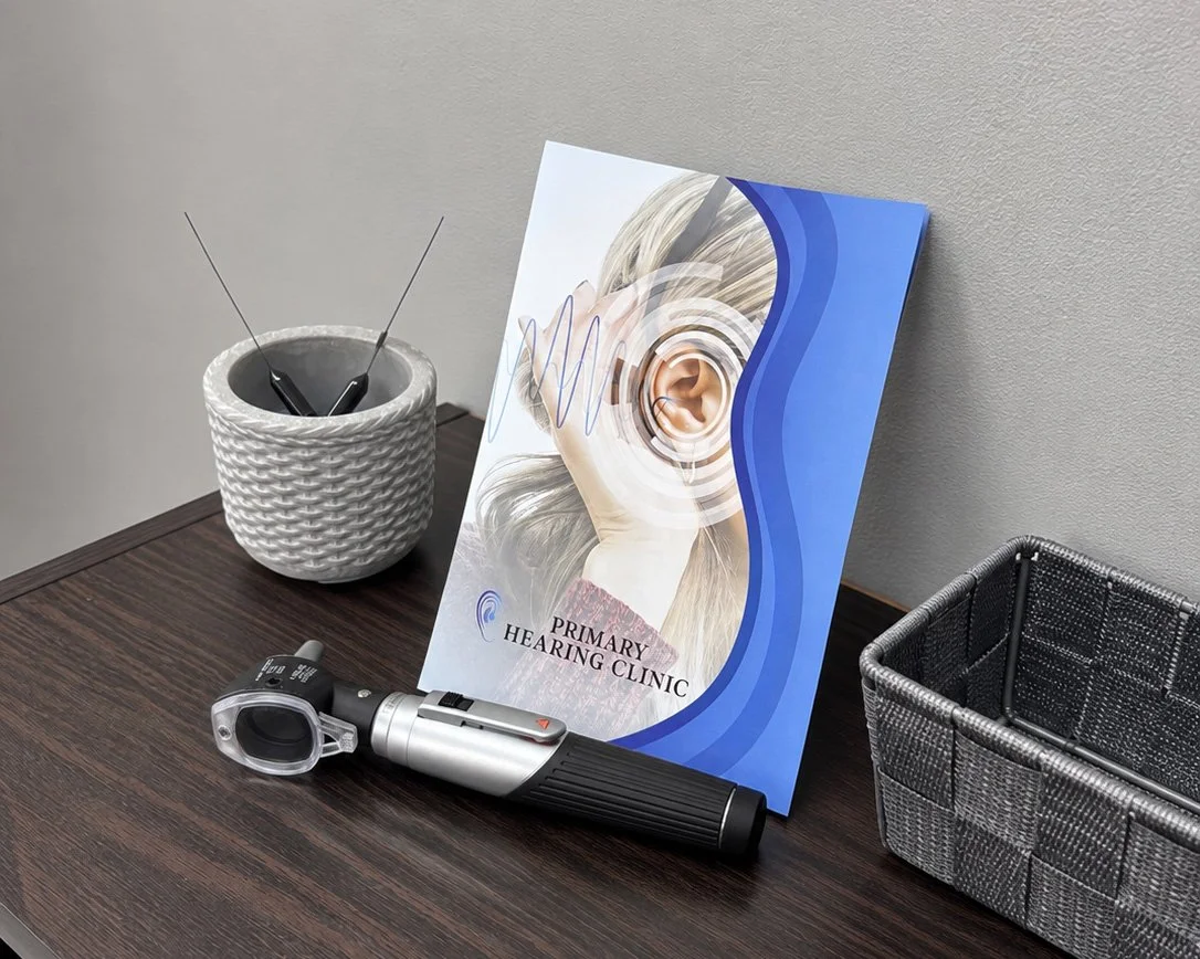 A brochure for a primary hearing clinic, a hearing test device, a gray flowerpot with two probes, and an empty gray woven basket on a dark wooden desk against a beige wall.
