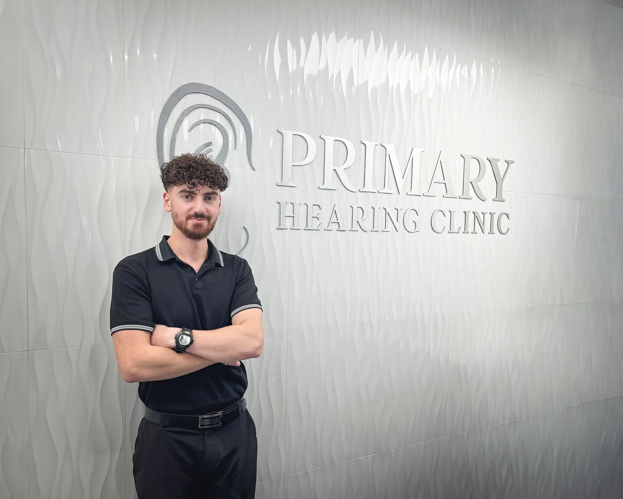 A man with curly dark hair and a beard crossing his arms, standing in front of a white, textured wall with the words 'PRIMARY HEARING CLINIC' and a logo. He is wearing a black polo shirt and a wristwatch.