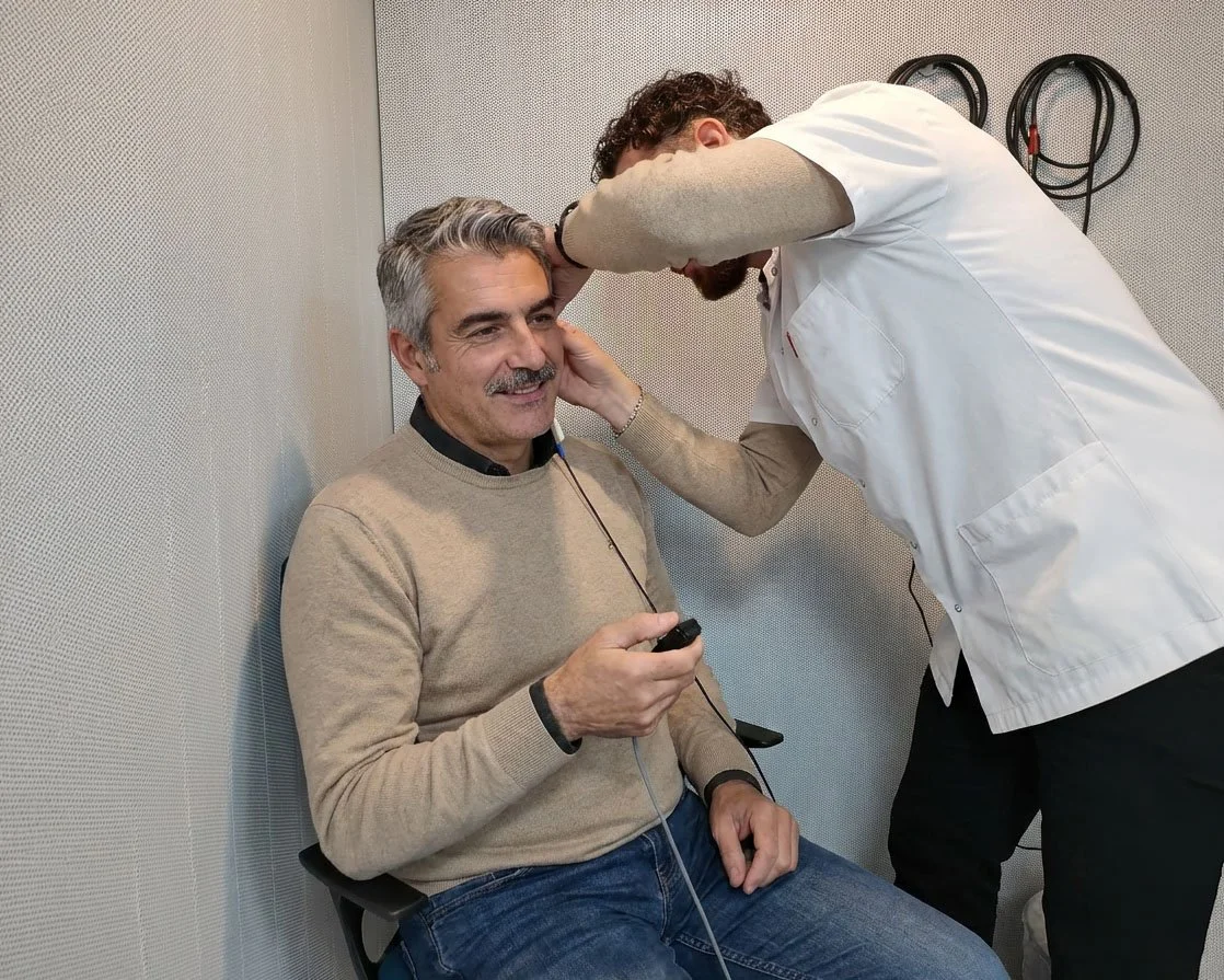 A man sitting in a chair, wearing headphones, smiling as another person adjusts equipment near his head in a clinical or testing setting.