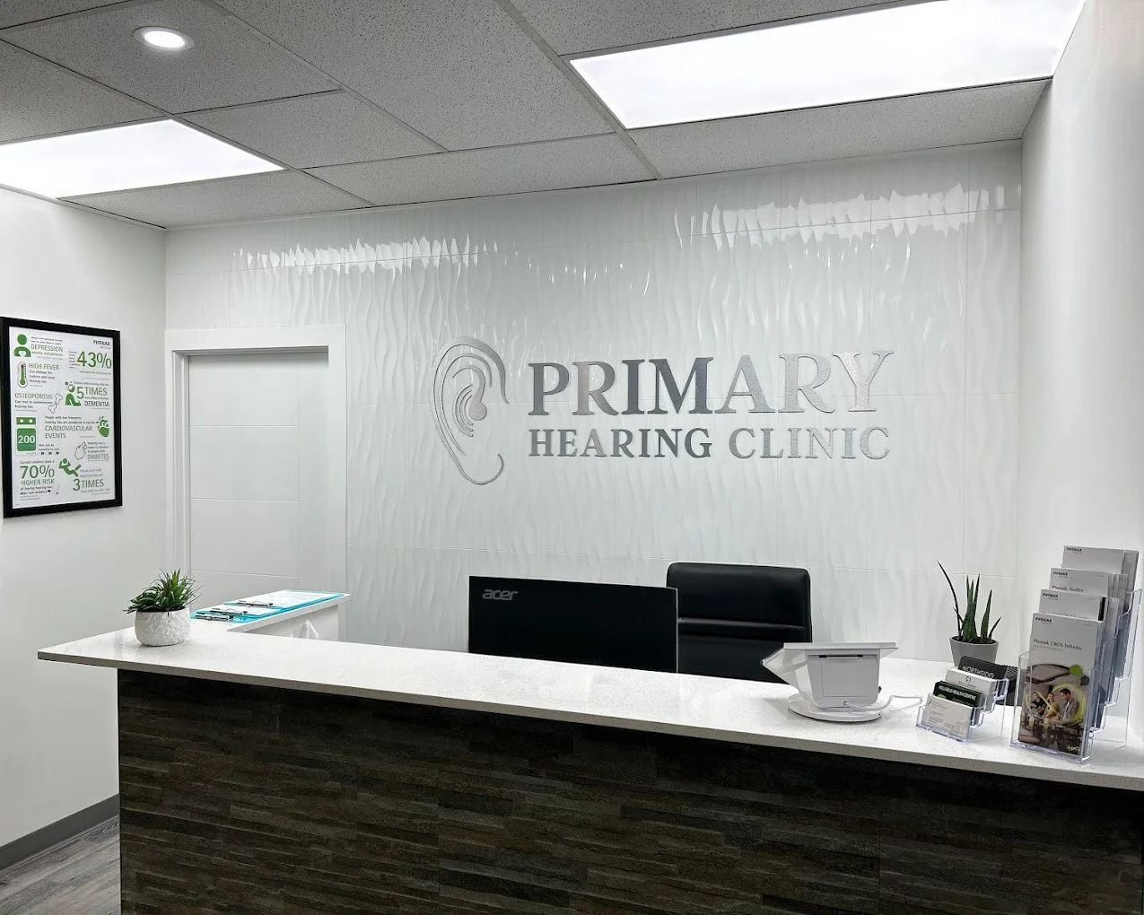Reception desk at Primary Hearing Clinic with PC monitor, chairs, potted plants, brochures, and clinic sign on wall.