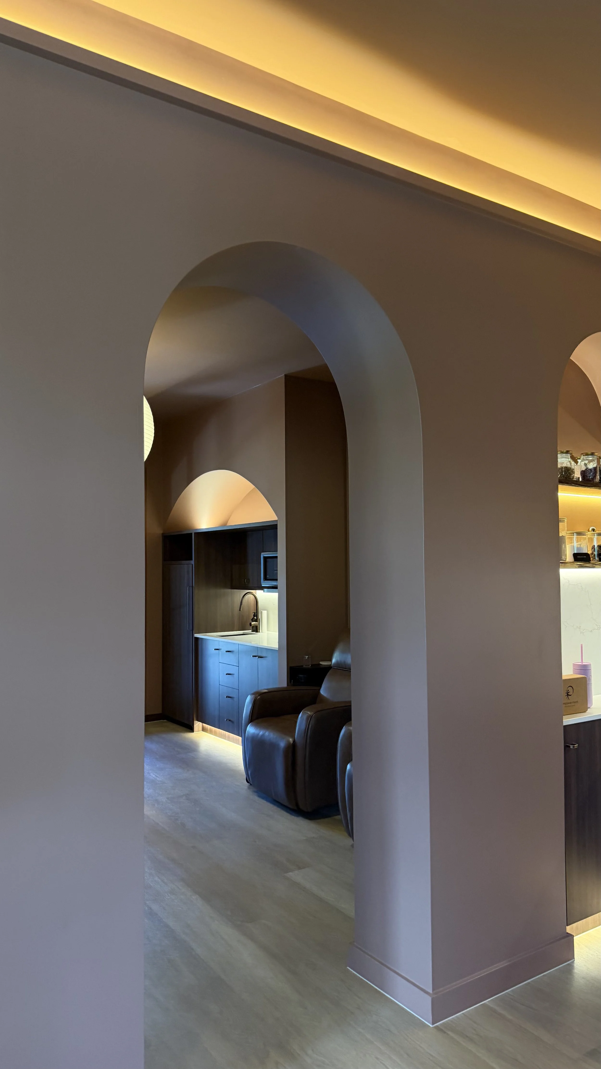 Interior of a modern living space with arched doorways, a small kitchen area with dark wood cabinets, white countertops, a sink, and a microwave. There are comfortable brown leather armchairs and soft warm lighting.