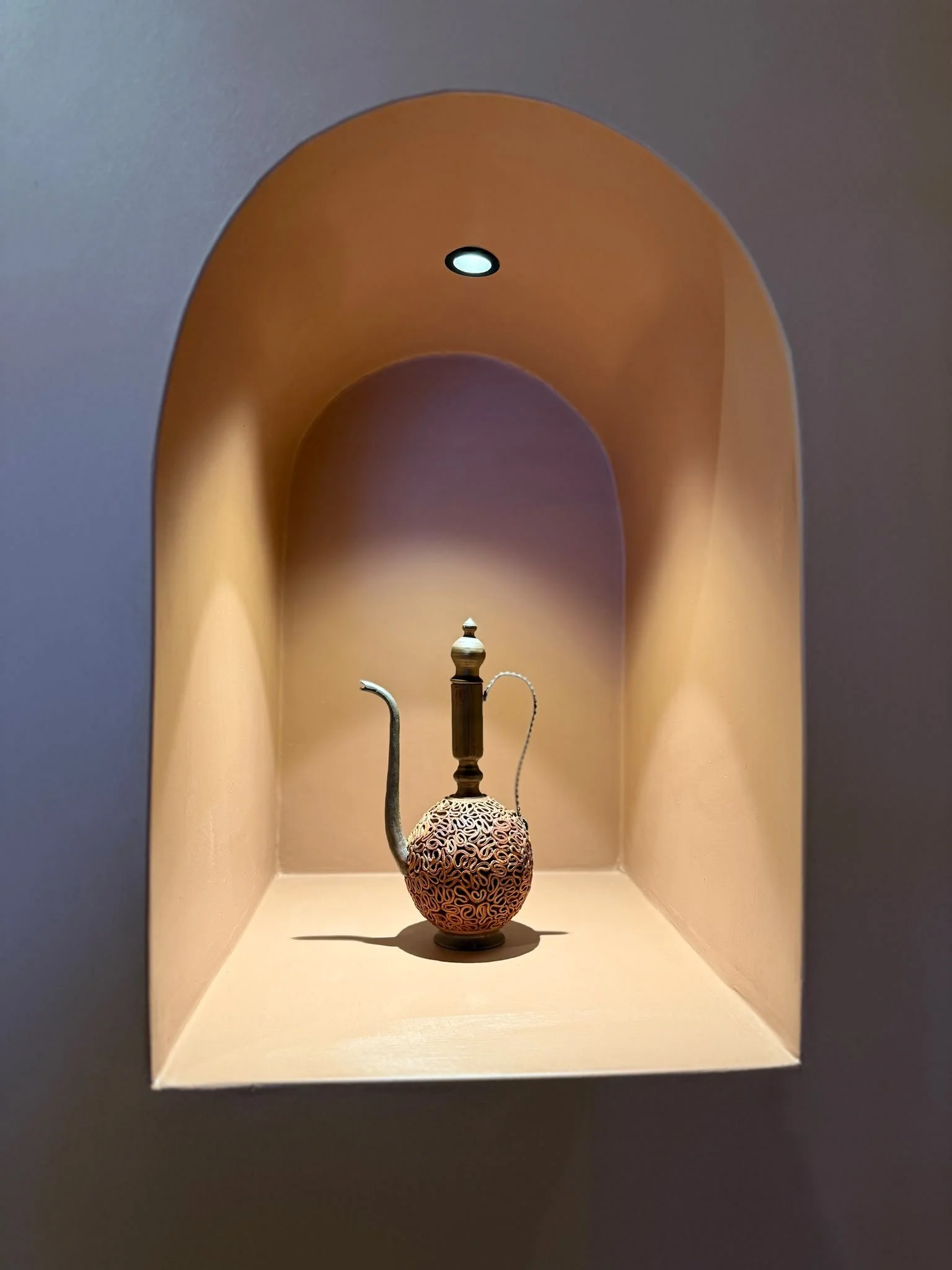 An ornate teapot with a decorative lid and metal spout, displayed in a niche with warm lighting.