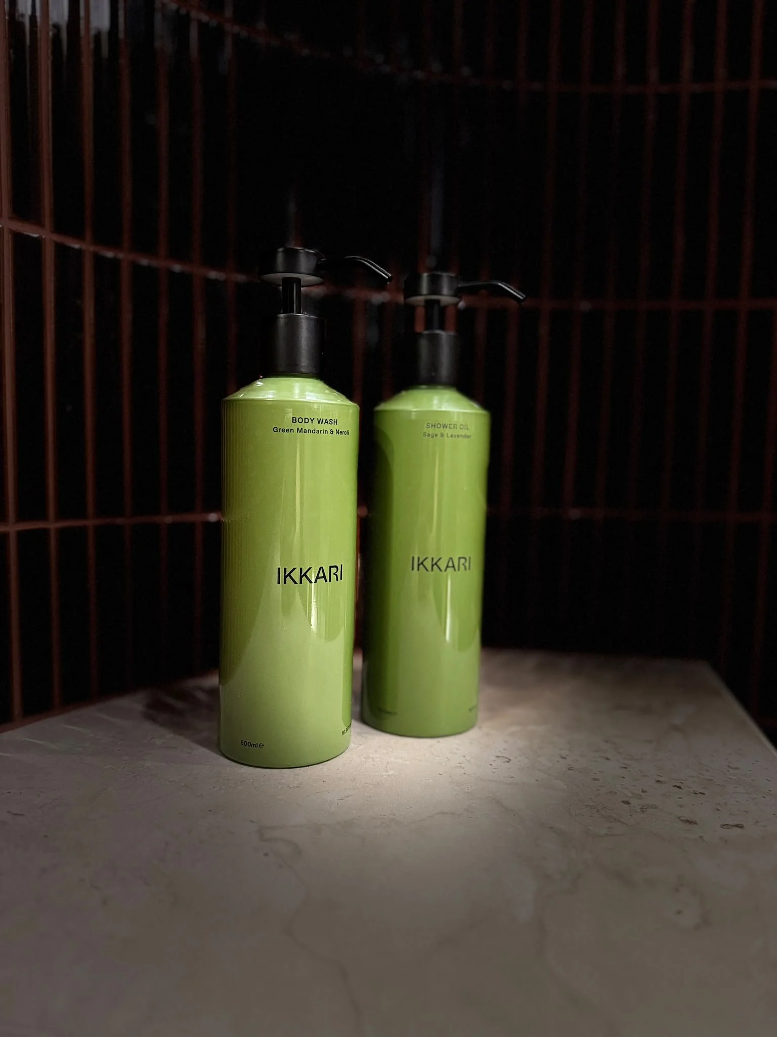Two green bottles labeled 'Body Wash Green Mandarin & Neroli' and 'Shower Oil Sage & Lavender' are placed on a gray surface inside a dark, grid-like enclosure with red metal bars. Ikkari bath and products. organic. health conscious