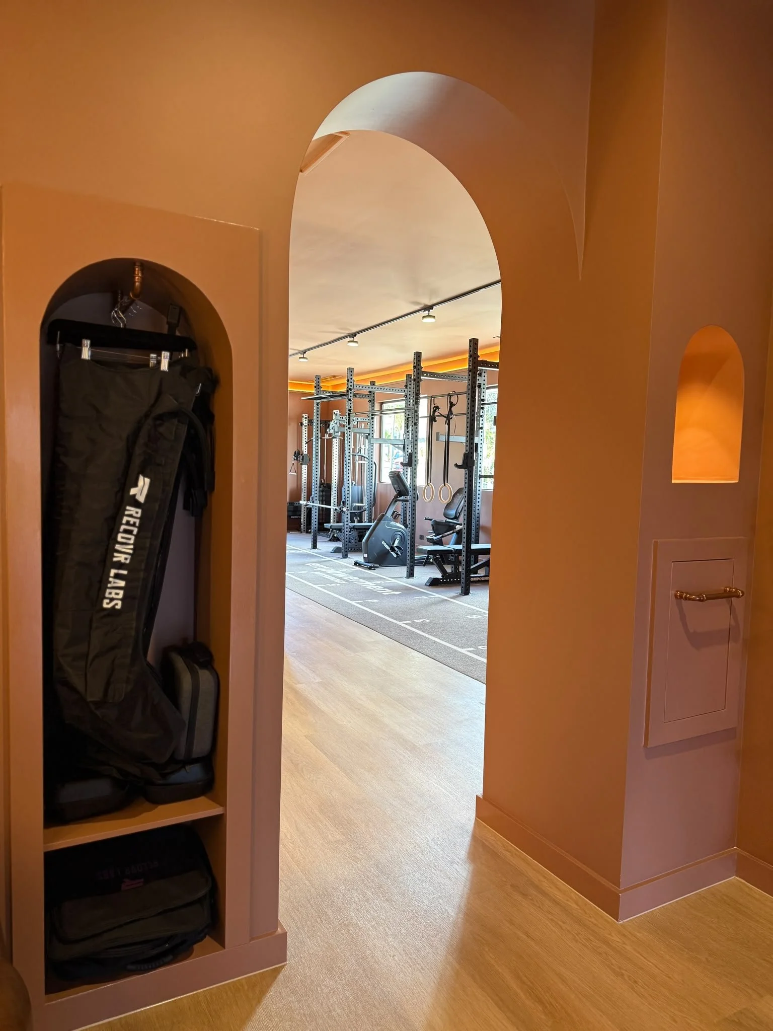 View of a gym with workout equipment seen through a decorative framed archway, featuring a Pilates mat and backpacks on shelves in the foreground.