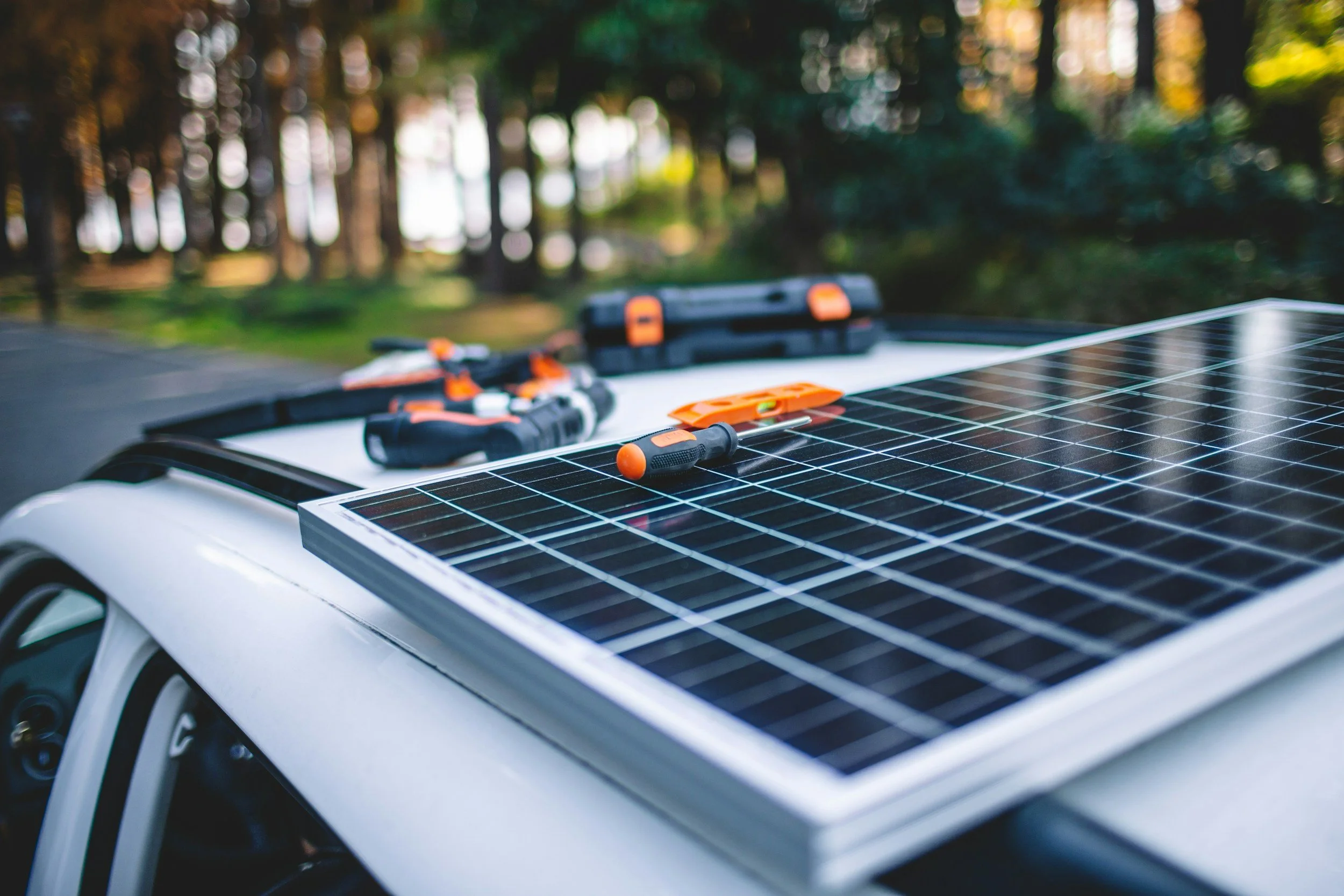 A solar panel mounted on the hood of a white vehicle outdoors, with tools and equipment placed on the panel, and a background of trees and sunlight filtering through.