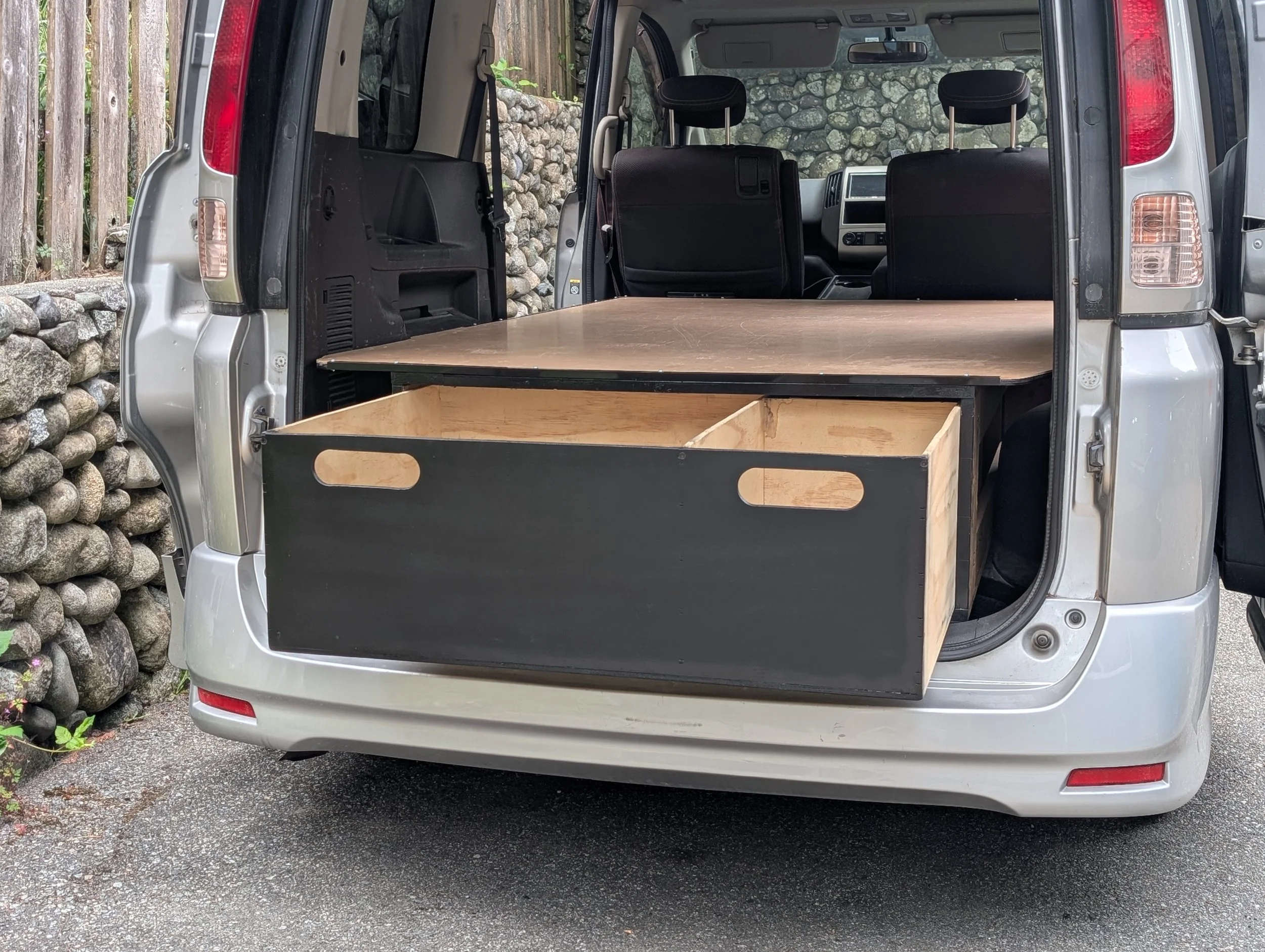The image shows the open back of a silver van with a wooden platform and storage drawer inside. The van is parked outdoors near a stone wall and a wooden fence.