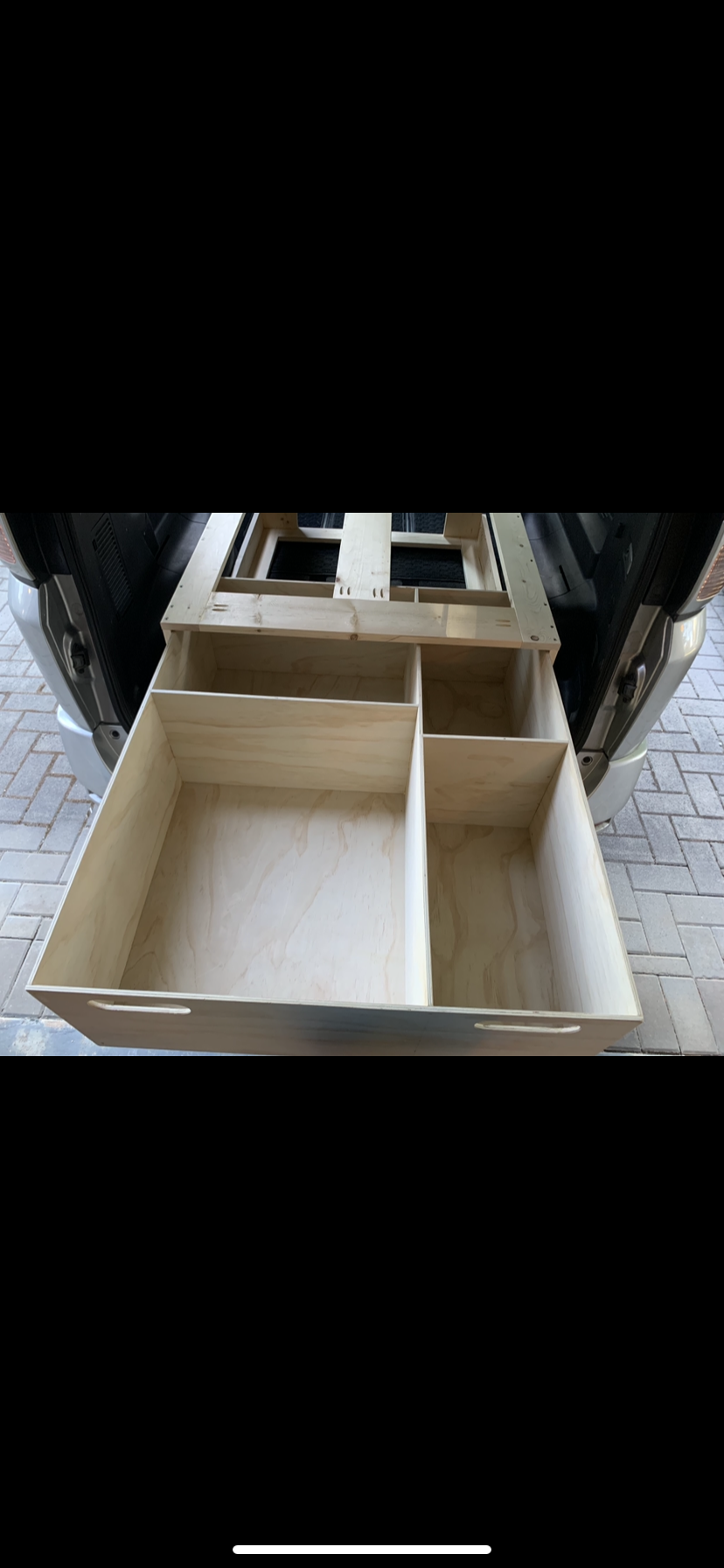Empty wooden drawer with multiple compartments placed inside an open vehicle trunk, on a brick-paved surface.