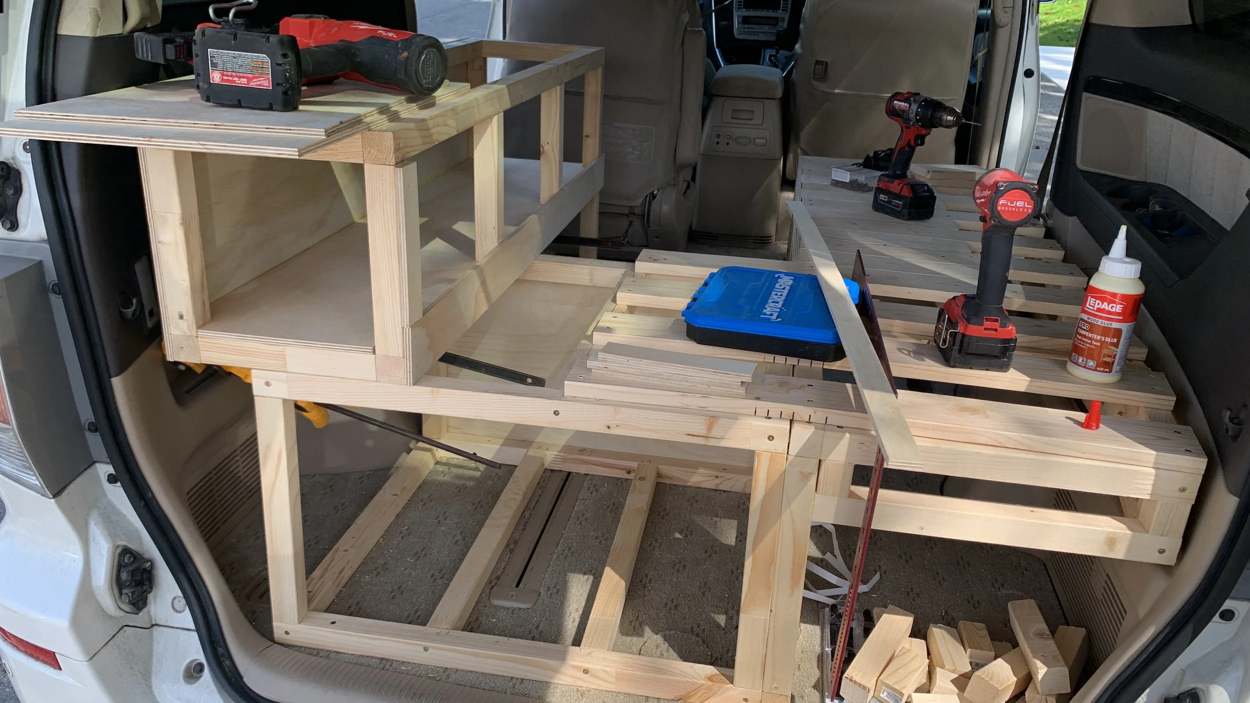 The image shows the interior of a vehicle with a partially built wooden furniture frame on a workbench, surrounded by tools including a cordless drill, a bottle of wood glue, and a blue storage container.