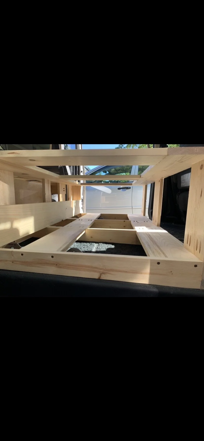 A wooden cube-shaped structure under construction, built with unfinished light-colored wood, placed on a car seat near a door and a van visible through the window in the background.