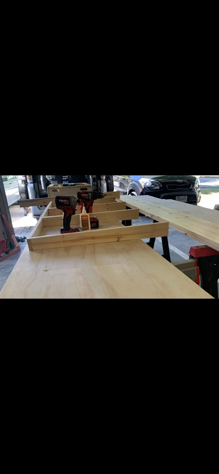 Woodworking workshop with a workbench, power drills, and wooden planks, with cars parked outside in the background.