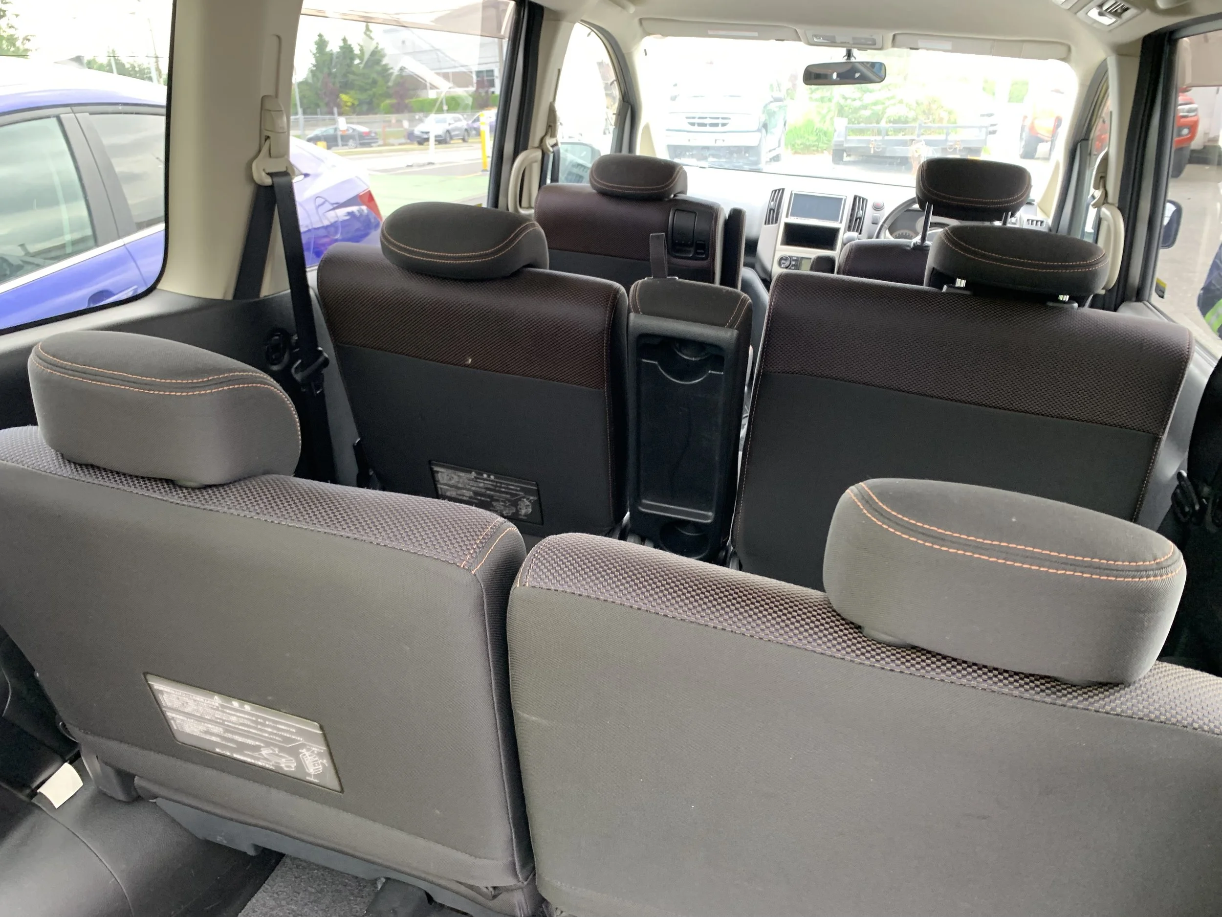Interior of a vehicle showing multiple rows of black fabric seats with brown stitching, some headrests, and large front windshield revealing a parking lot with several cars and trees outside.