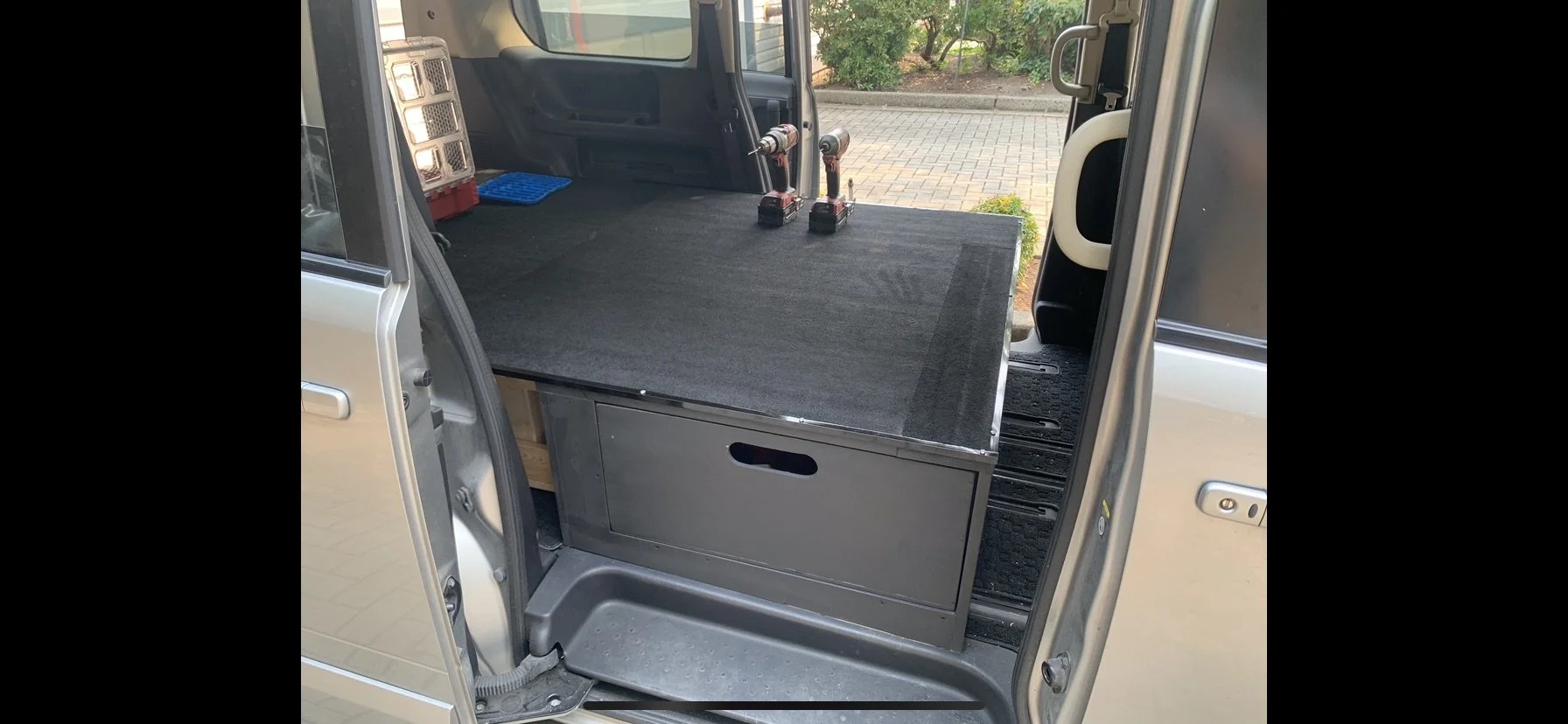 Inside a van with a raised platform or storage area, two cordless drills, a blue ice pack, and a small red crate with compartments, with a view of a paved sidewalk and greenery outside.