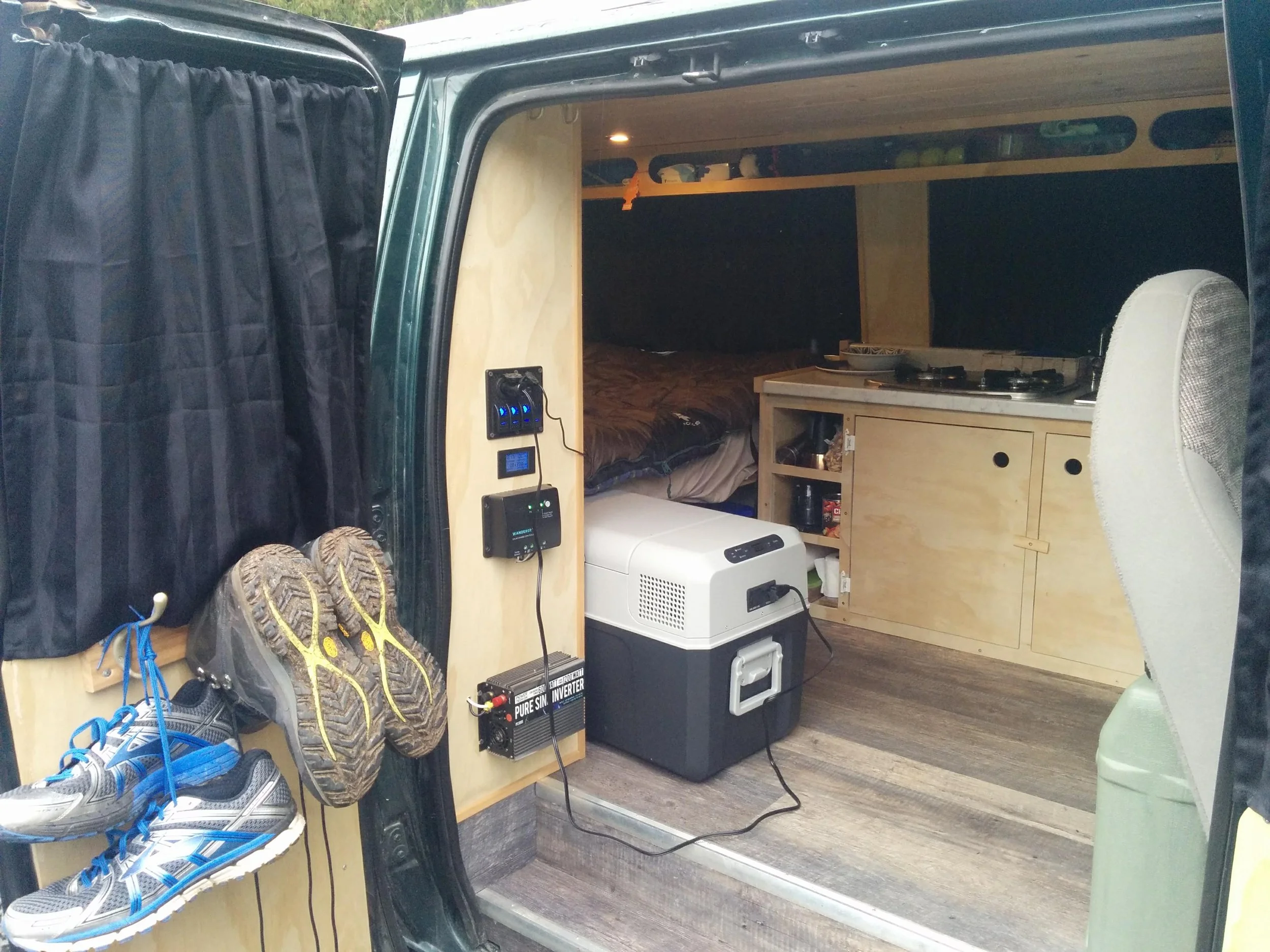 Interior of a small camper van with wood-paneled walls, a bed, a small kitchenette, and shelves with green storage containers. Shoes and outdoor gear are near the entrance, and a power panel and small inverter are mounted on a wood panel.