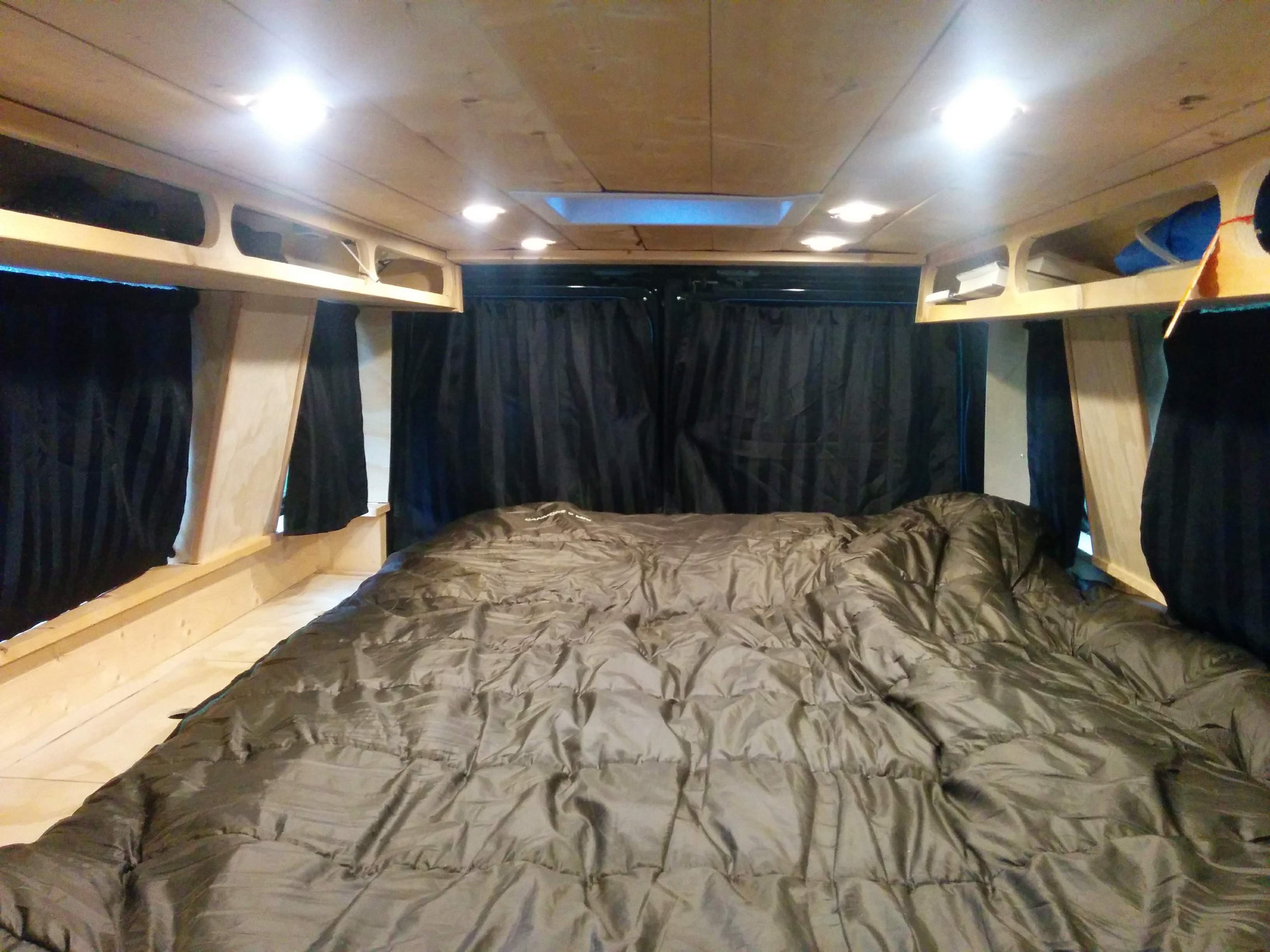 Interior of a small camper or RV with a bed, wooden overhead storage compartments, and black curtains.