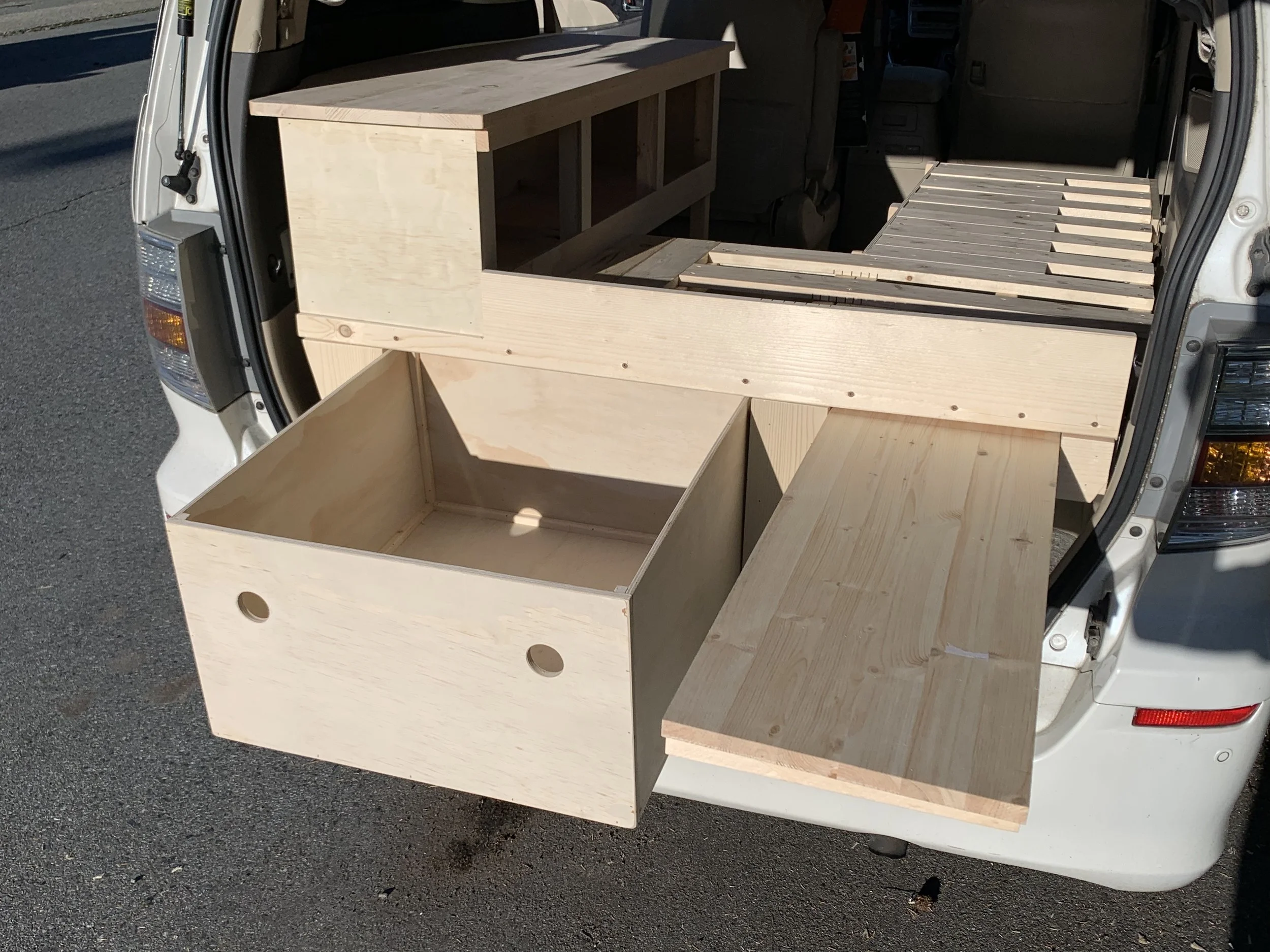 The back of a white van transporting unfinished wooden furniture, including a partially assembled cabinet and slatted furniture pieces, parked on an asphalt street.