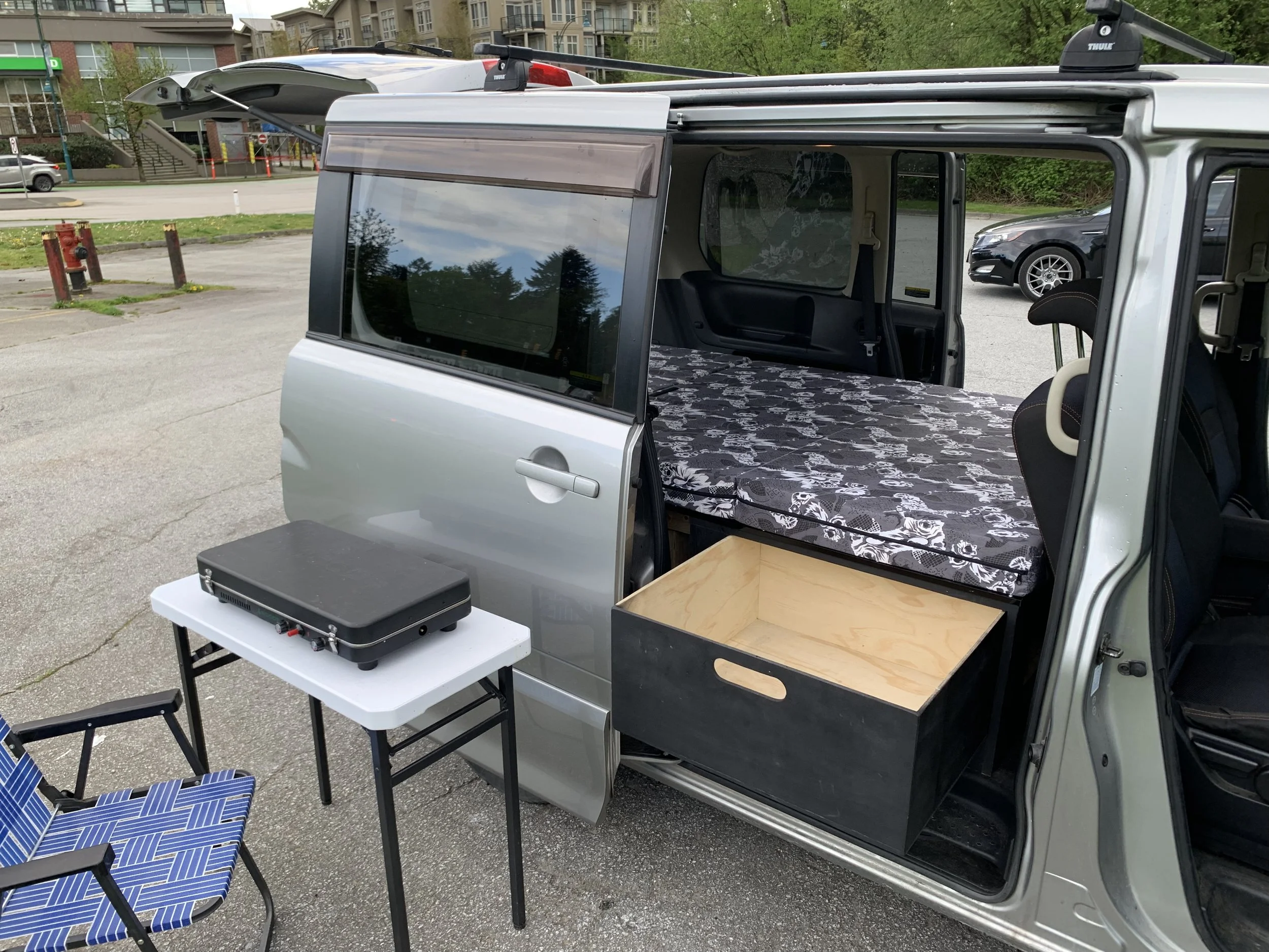 A van with its side door open, acting as a mobile workspace or setup for an outdoor event. Inside, there is a patterned mattress, a table with a portable stove, and some chairs outside the van.