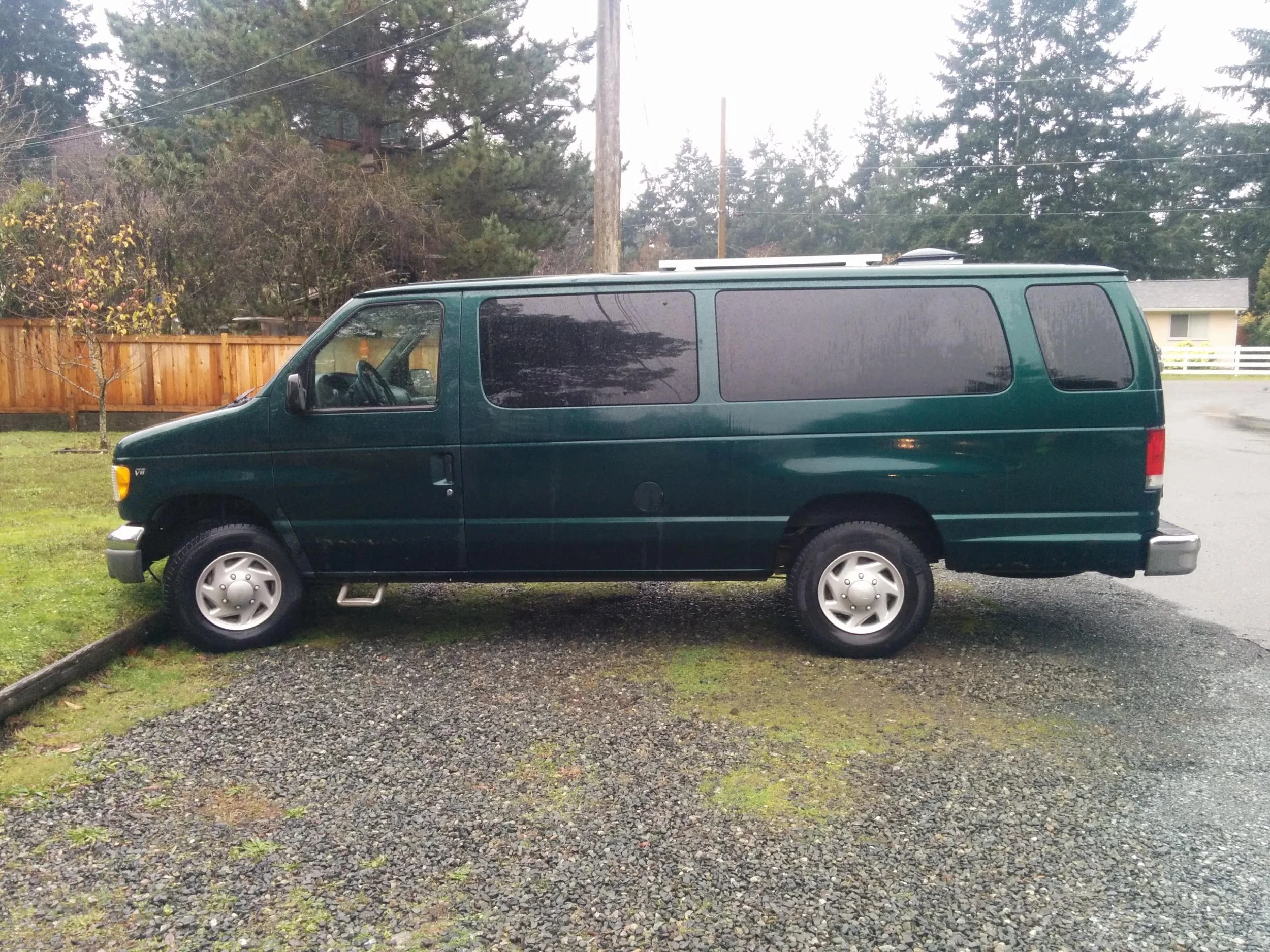 A green van parked on a gravel driveway next to a grassy area with a small tree. In the background, there are tall trees, a wooden fence, and a house with a white fence. The sky appears overcast.
