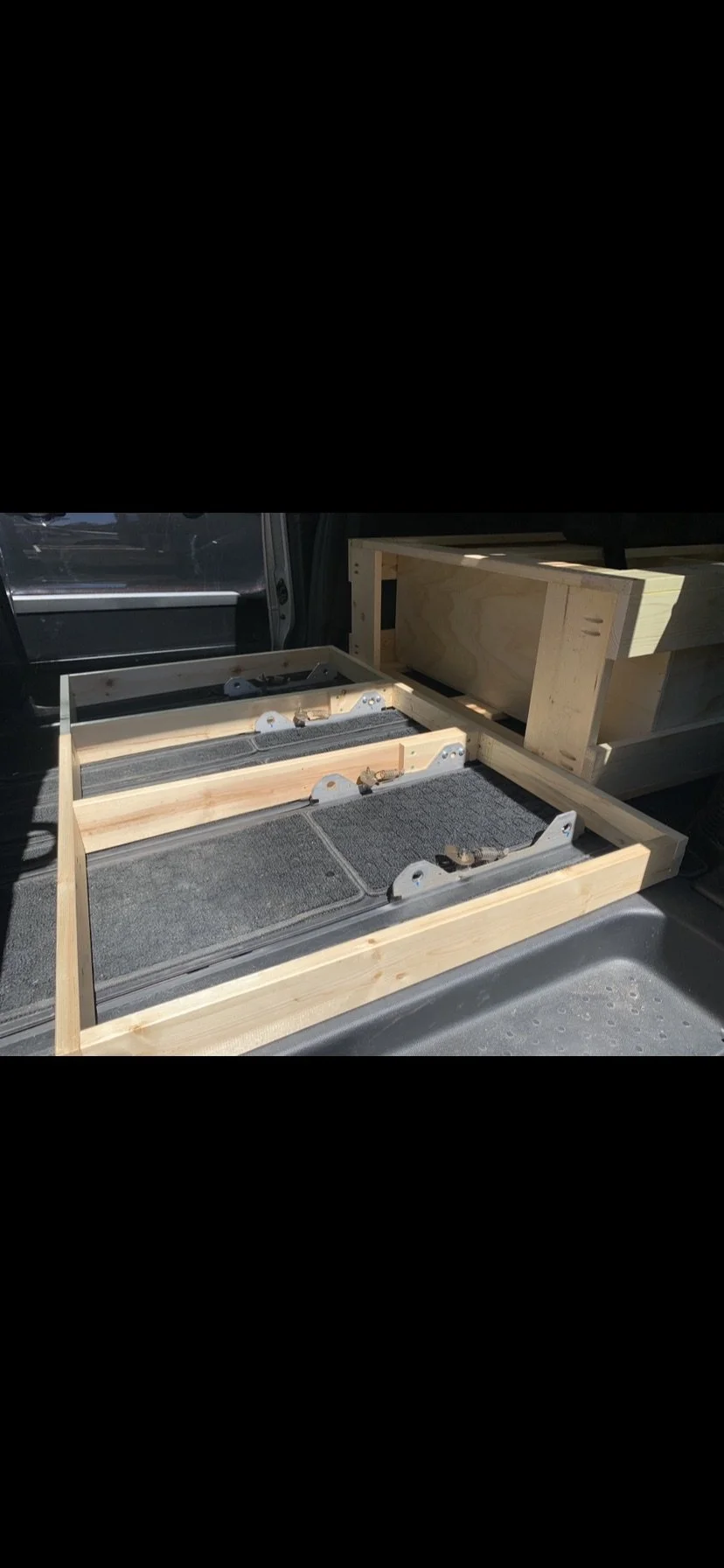 Wooden crate with metal brackets, placed on the floor of a vehicle, with another partially assembled wooden structure behind it.