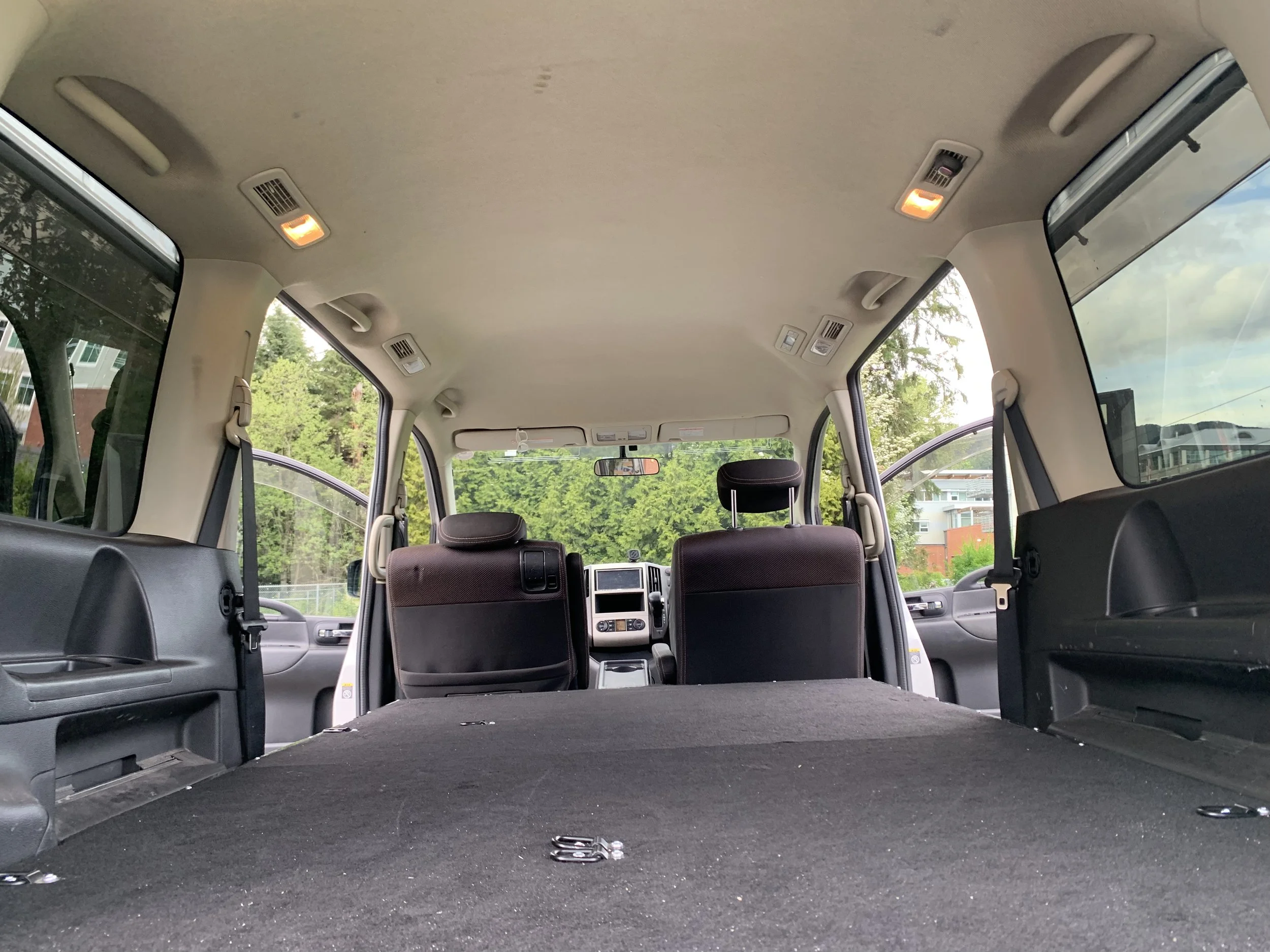 Inside the empty back of a vehicle, showing the rear seats, headrests, and a flat cargo area with some clips and hooks, with windows revealing trees and nearby houses.