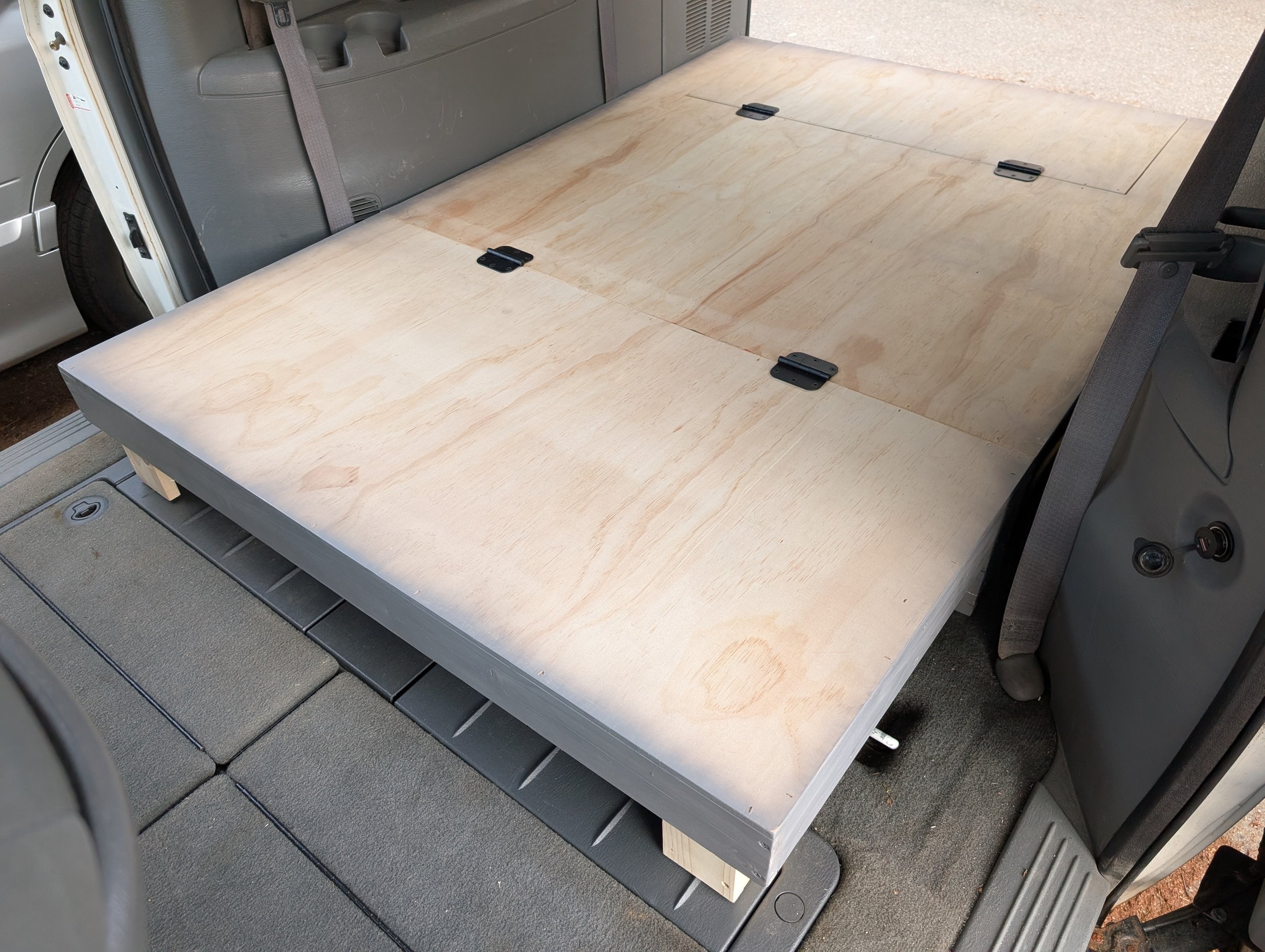 The interior of a vehicle showing the cargo area with a flat plywood platform and metal hinges, possibly for custom storage or conversion.