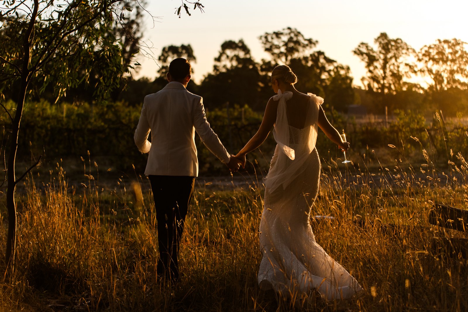 A newly married couple walking hand in hand through long grass at sunset in regional Australia. Storytelling wedding photography by Georgie James Photography.