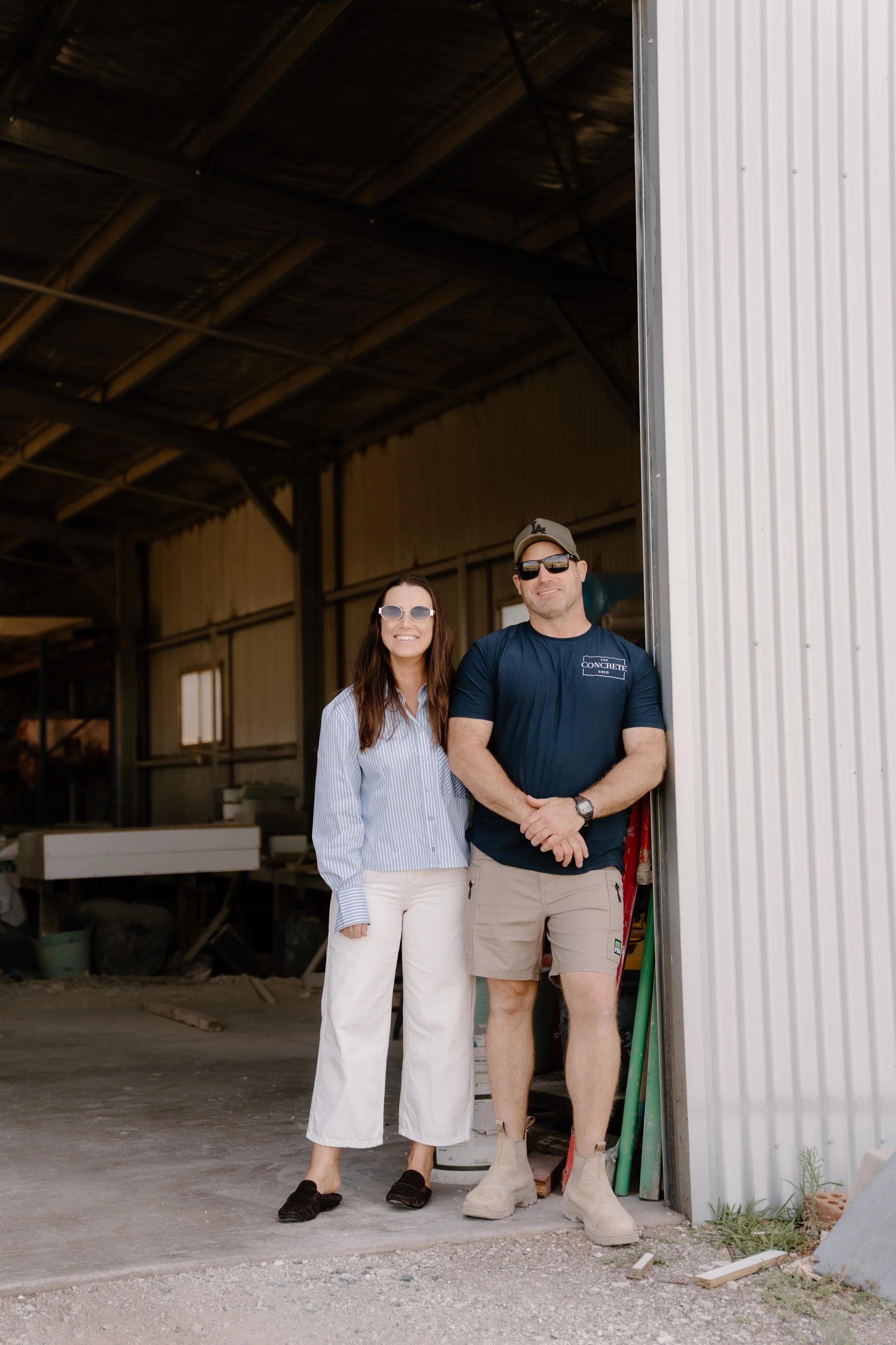 A woman and a man stand side by side in front of an open garage door with a construction site visible inside, smiling at the camera. The woman wears sunglasses, a striped shirt, and white pants, while the man wears sunglasses, a baseball cap, a navy T-shirt, and beige shorts.