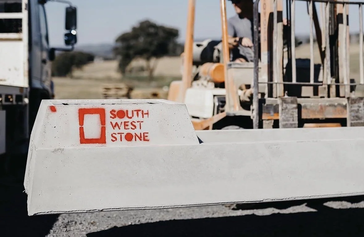 A white concrete block with red spray paint that reads 'South West Stone' and a red square outline, in an outdoor stone yard with equipment and a person in the background.