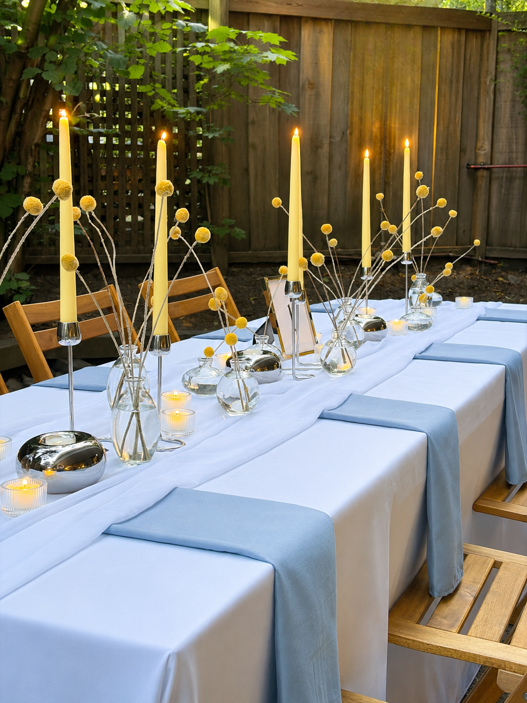 A long outdoor dining table decorated with white tablecloths, light blue napkins, tall yellow candles in glass holders, small vases with yellow billy balls, and small candles, set in a backyard with a wooden fence and trees in the background.