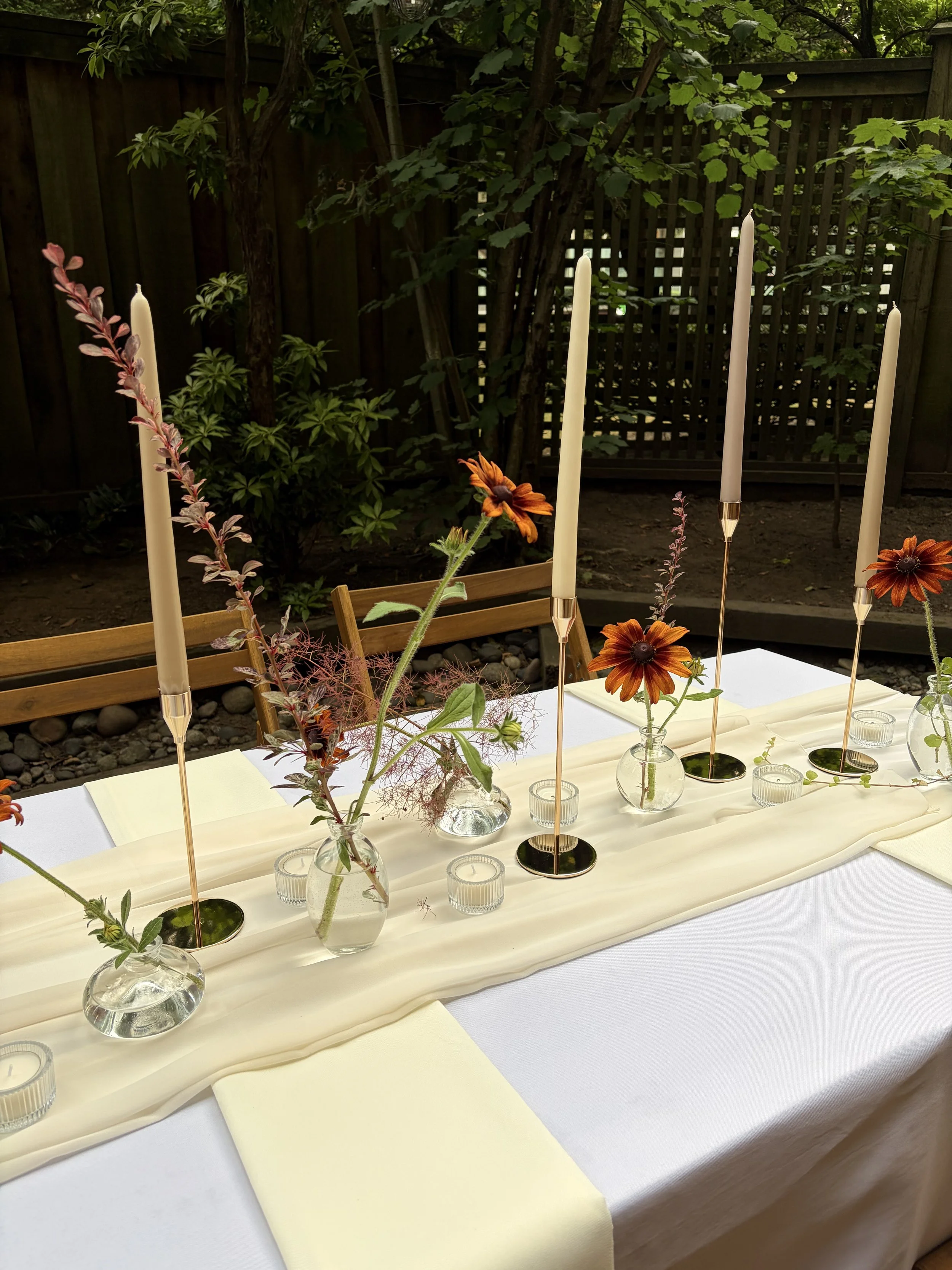 A dining table decorated with small vases holding flowers, tall candles, and candles in small glass holders, set outdoors on a white tablecloth with a yellow runner, surrounded by wooden chairs, with greenery and a wooden fence in the background.