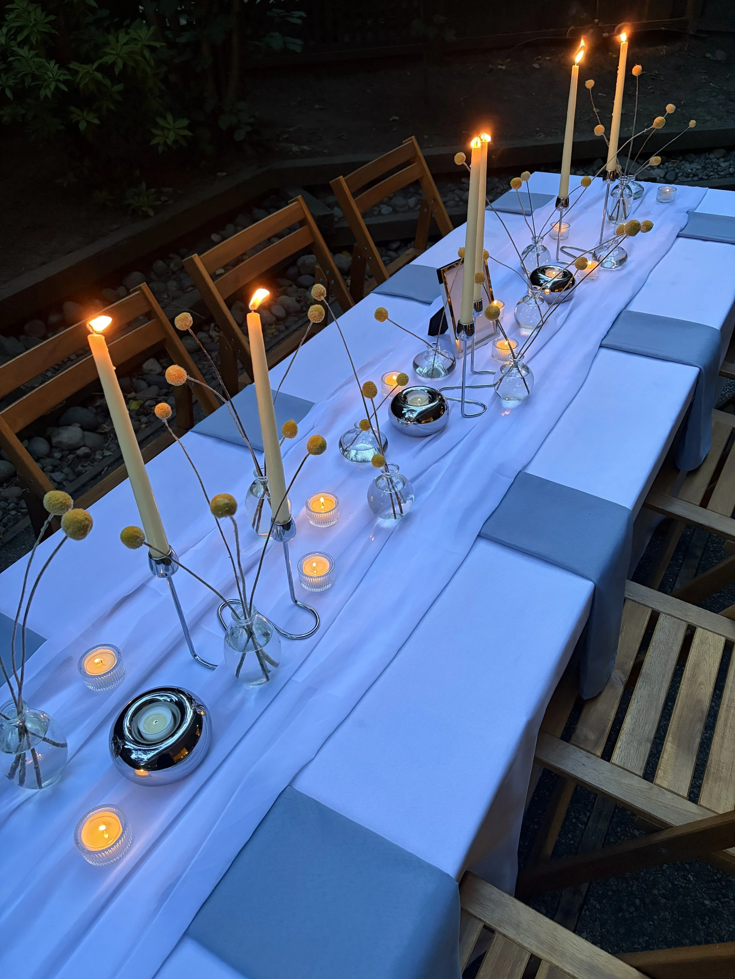 An outdoor dining table decorated with candles and yellow billy ball flowers in glass vases, set with blue napkins and white tablecloth, surrounded by wooden chairs, during evening time.