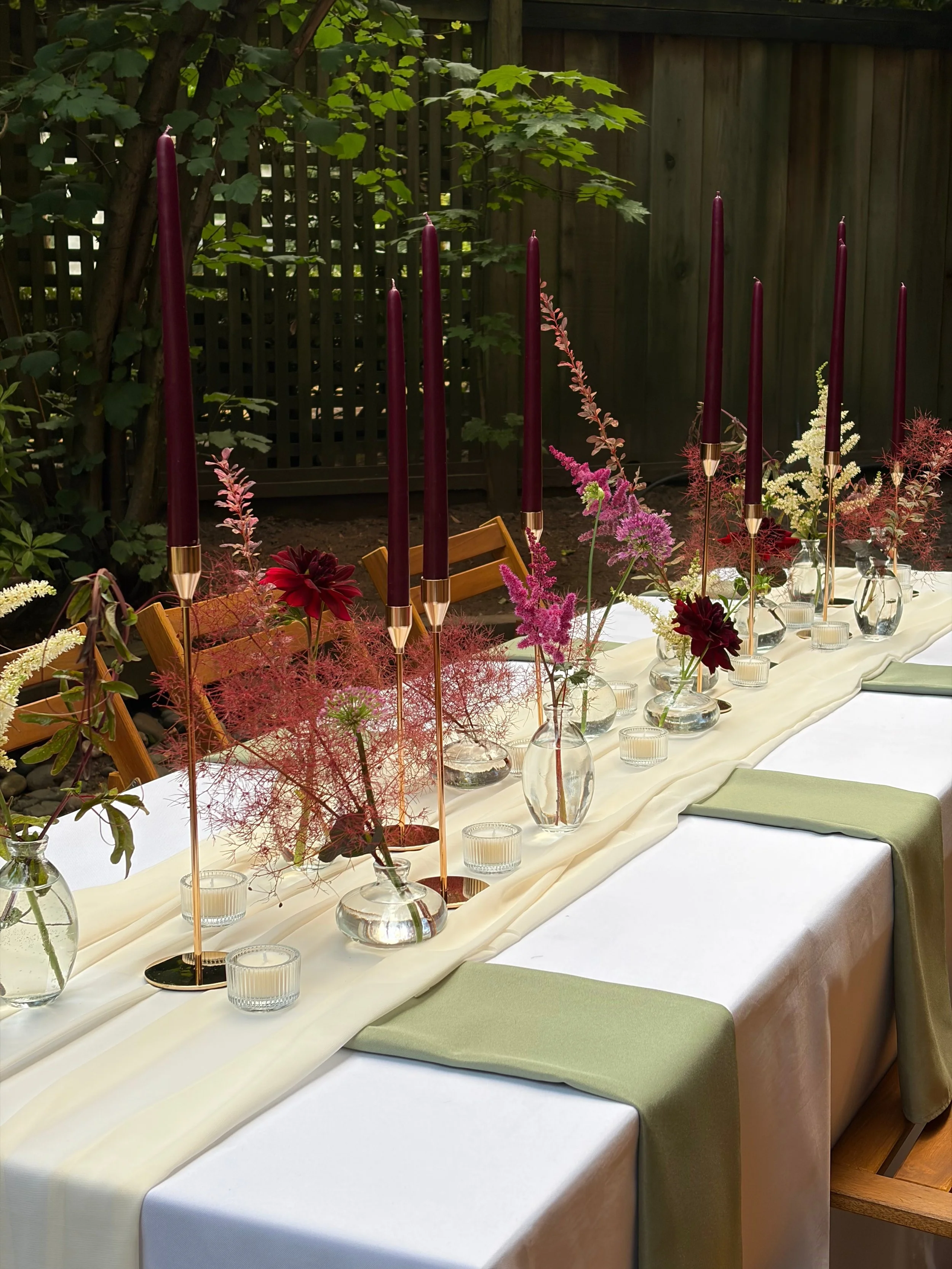 A decorated dining table with flowers in glass vases, tall dark purple candles, and small glass votive candles in a garden setting with a wooden fence and green foliage in the background.
