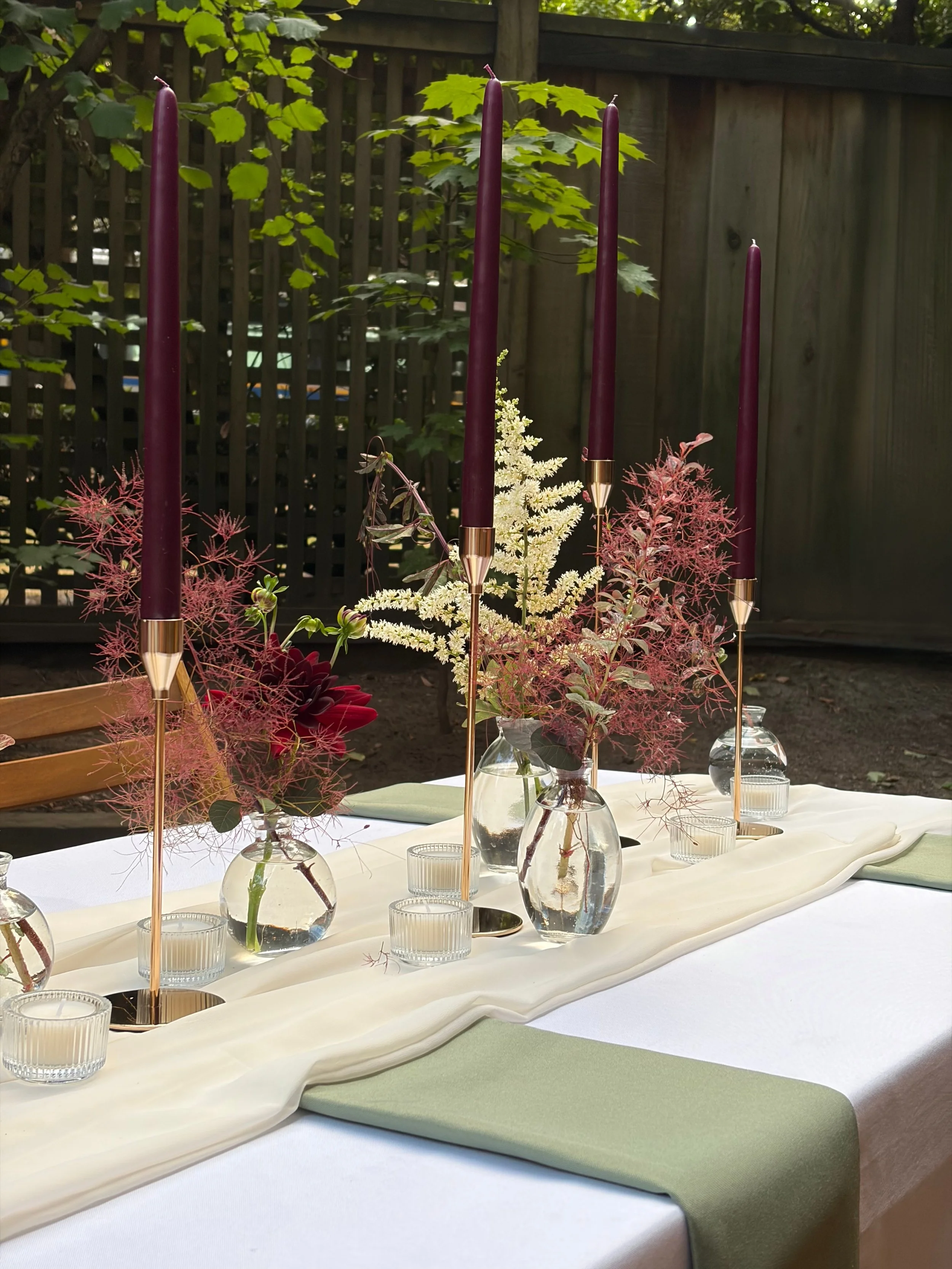 A table decorated with vases of flowers, tall purple candles in gold holders, and small glass candle holders, set outdoors with a white tablecloth and a green table runner, surrounded by a wooden fence and green foliage.
