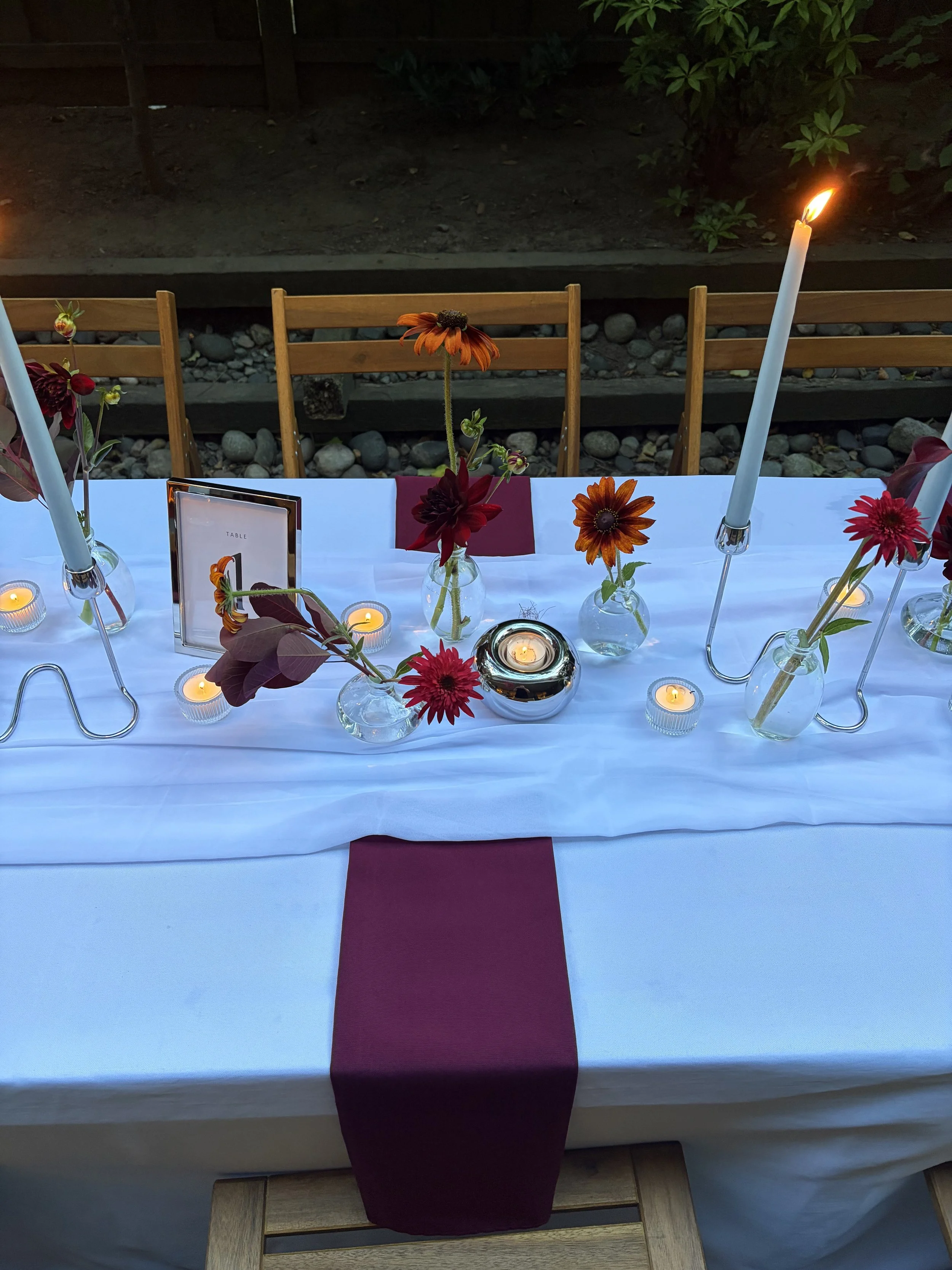 A table decorated for a dinner event with a white tablecloth, red napkin, small candles, and vases with orange and red flowers. There are wooden chairs behind the table, and the background features a dark outdoor area with a dirt ground, rocks, and g