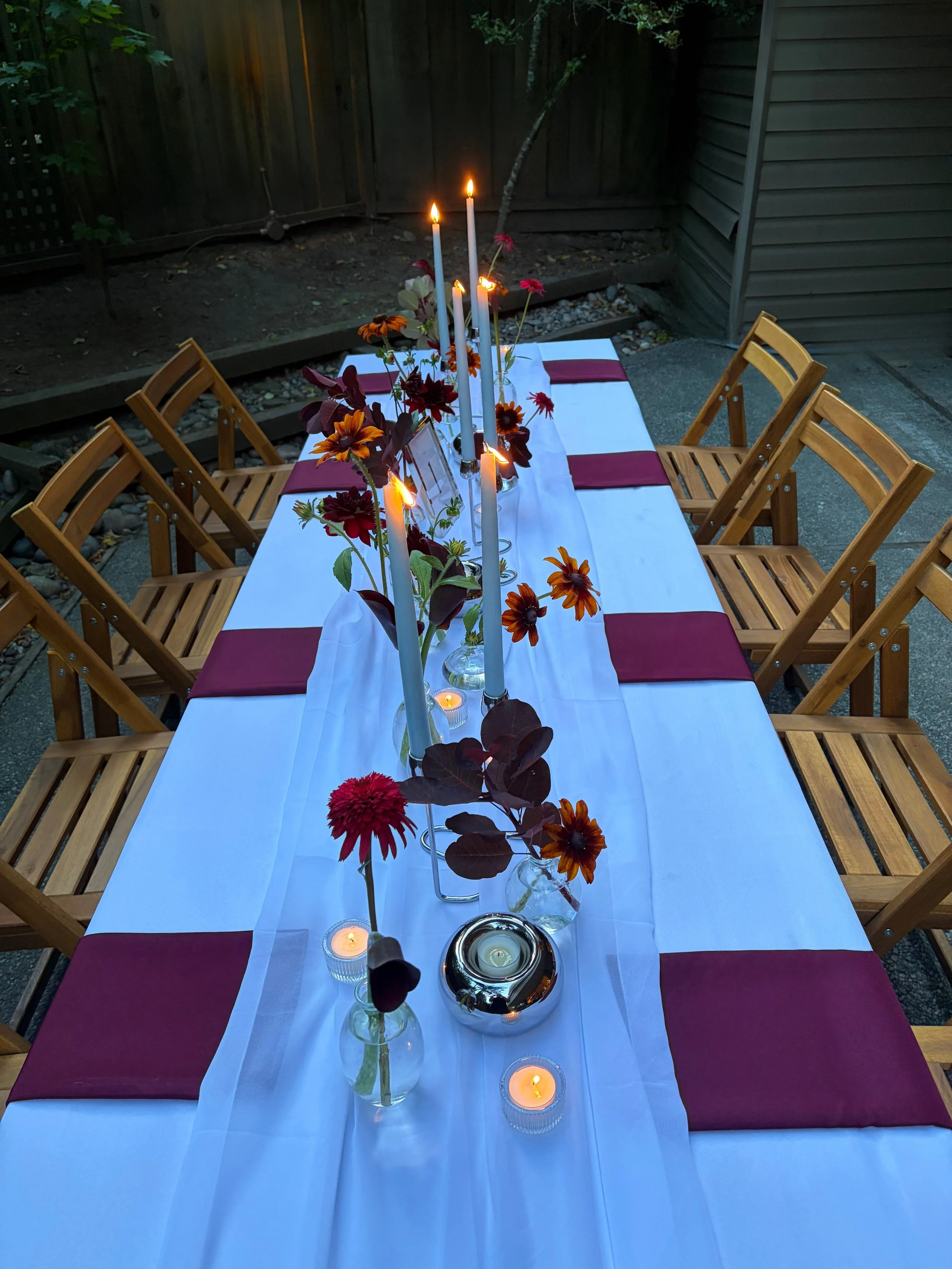 Outdoor dining table set for a celebration with a white tablecloth, red placemats, and a centerpiece of orange and red flowers. There are tall white candles and small tealight candles, some lit, creating a warm ambiance. Wooden chairs surround the ta