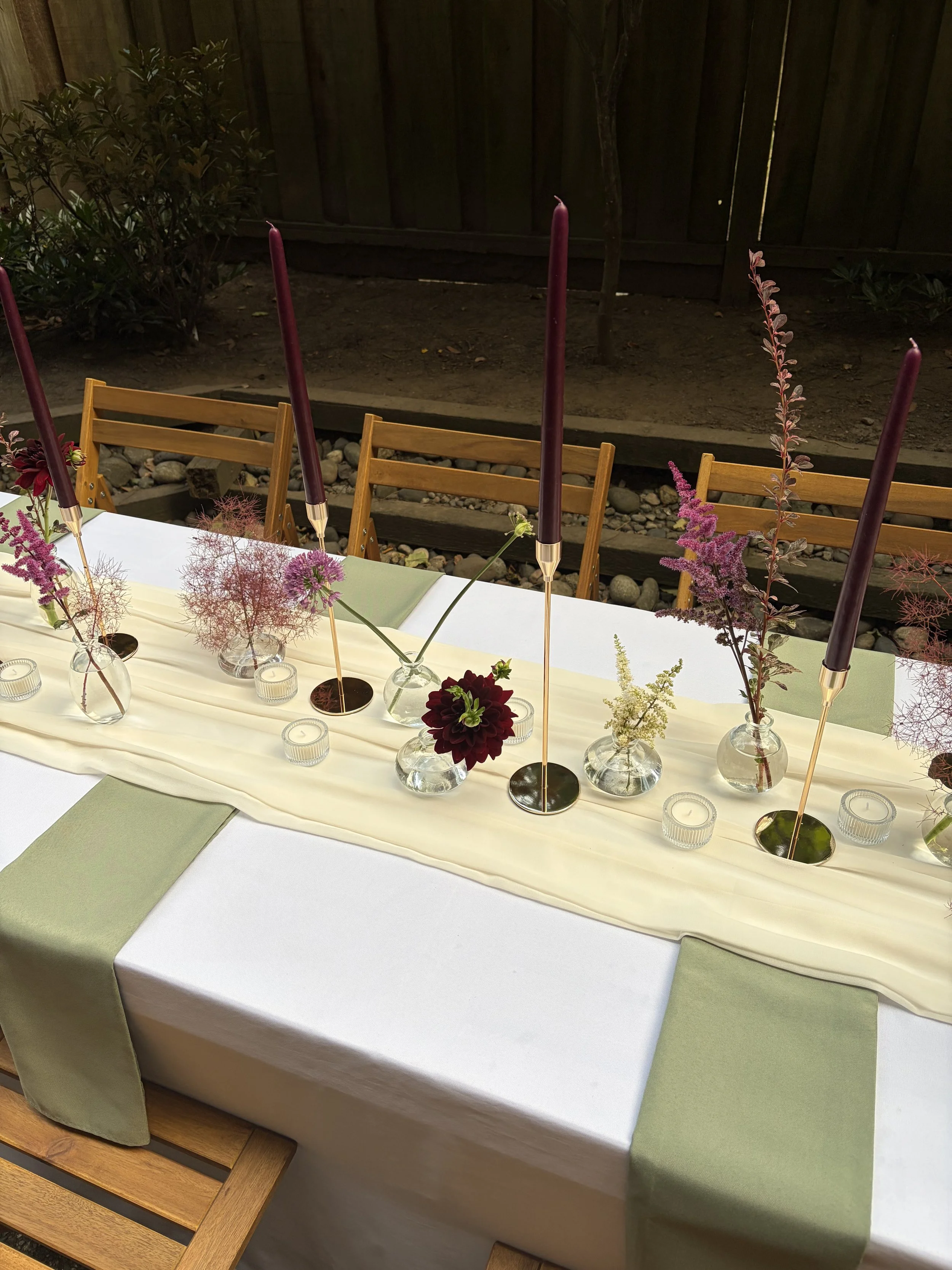 A long rectangular dining table with a white tablecloth and green placemats, decorated with tall purple candles in gold holders, clear glass vases with pink and purple flowers, and small glass candle holders, set outdoors against a wooden fence.
