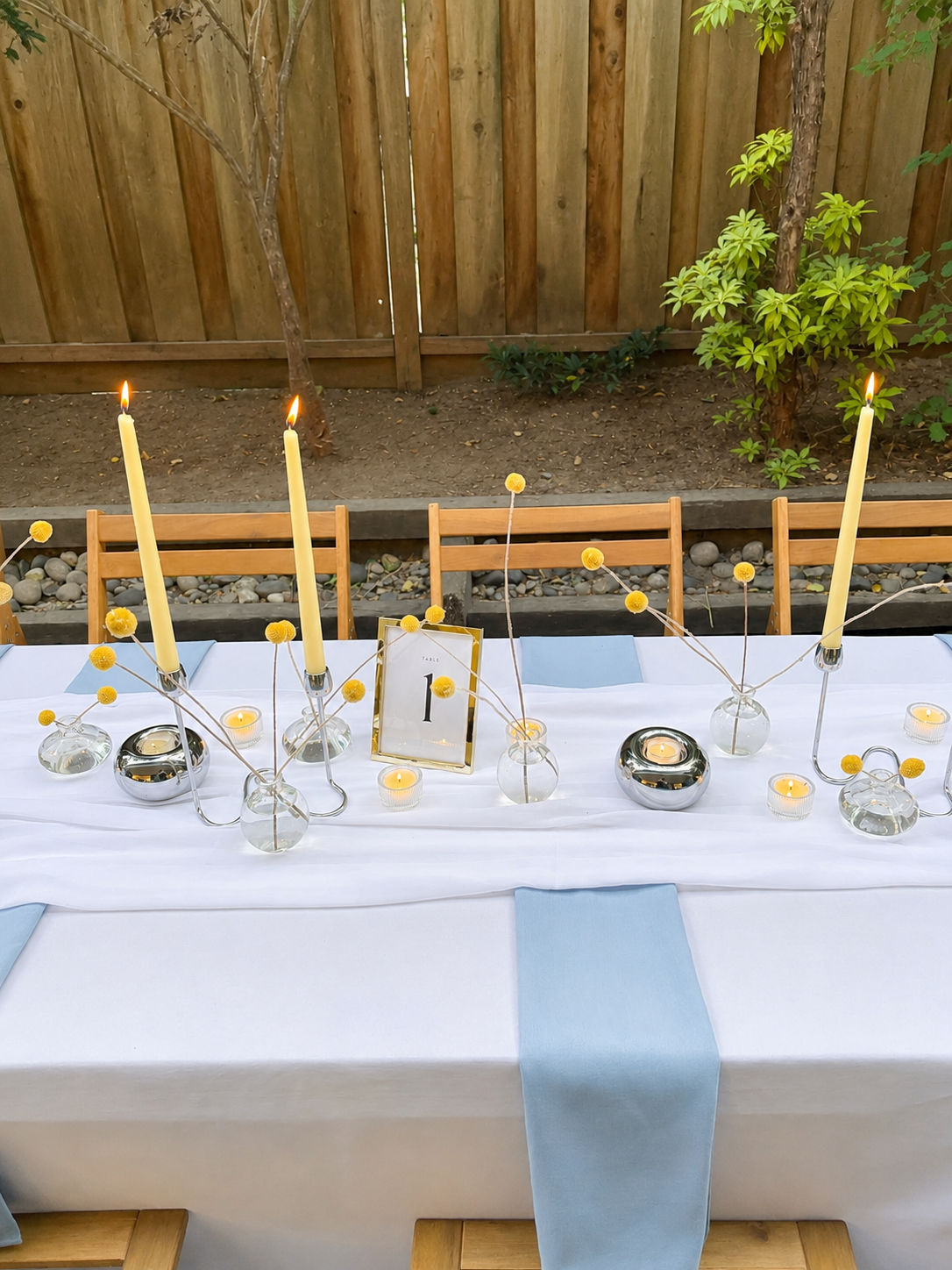 Elegant outdoor dining table with white tablecloth, light blue napkins, tall yellow candles, small tealight candles, glass vases with yellow pom-pom flowers, and a table number sign that reads 'Table 1'. A wooden fence and garden plants are in the ba