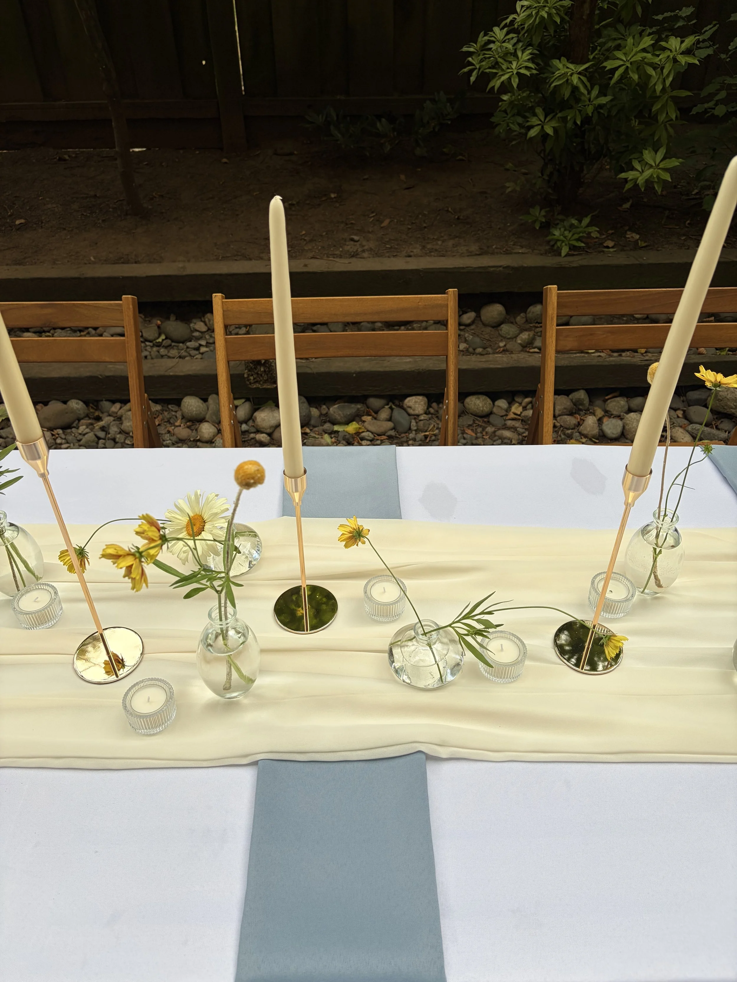 A table decorated with flowers, candles, and tall white candles in glass holders, with a light-colored tablecloth and a blue napkin on a chair. In the background, there are wooden chairs and a garden area with dirt, rocks, and green plants.