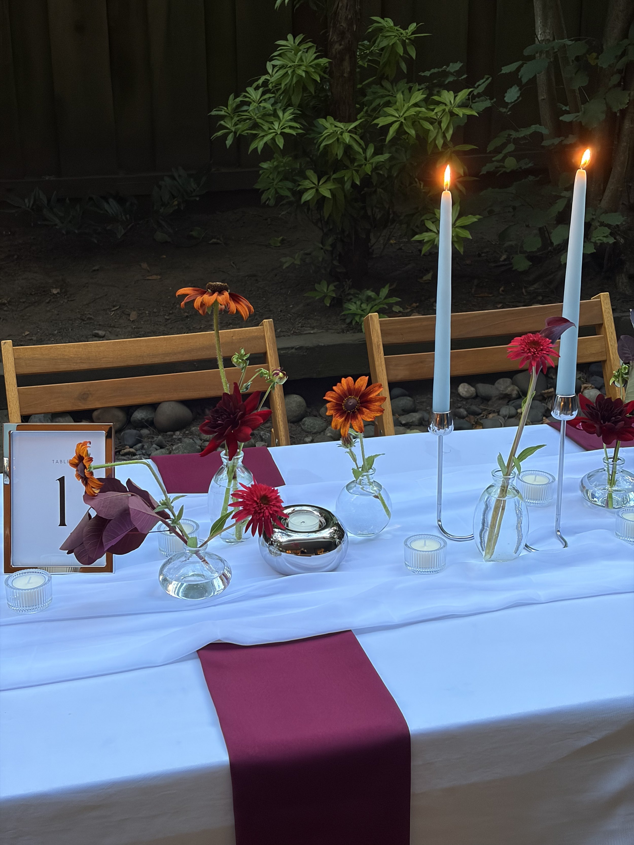 A table with a white tablecloth and a pink table runner, decorated with small glass vases of colorful flowers, and two tall blue candles burning. There is a table number card indicating it is table 1. The background shows wooden chairs and a garden w