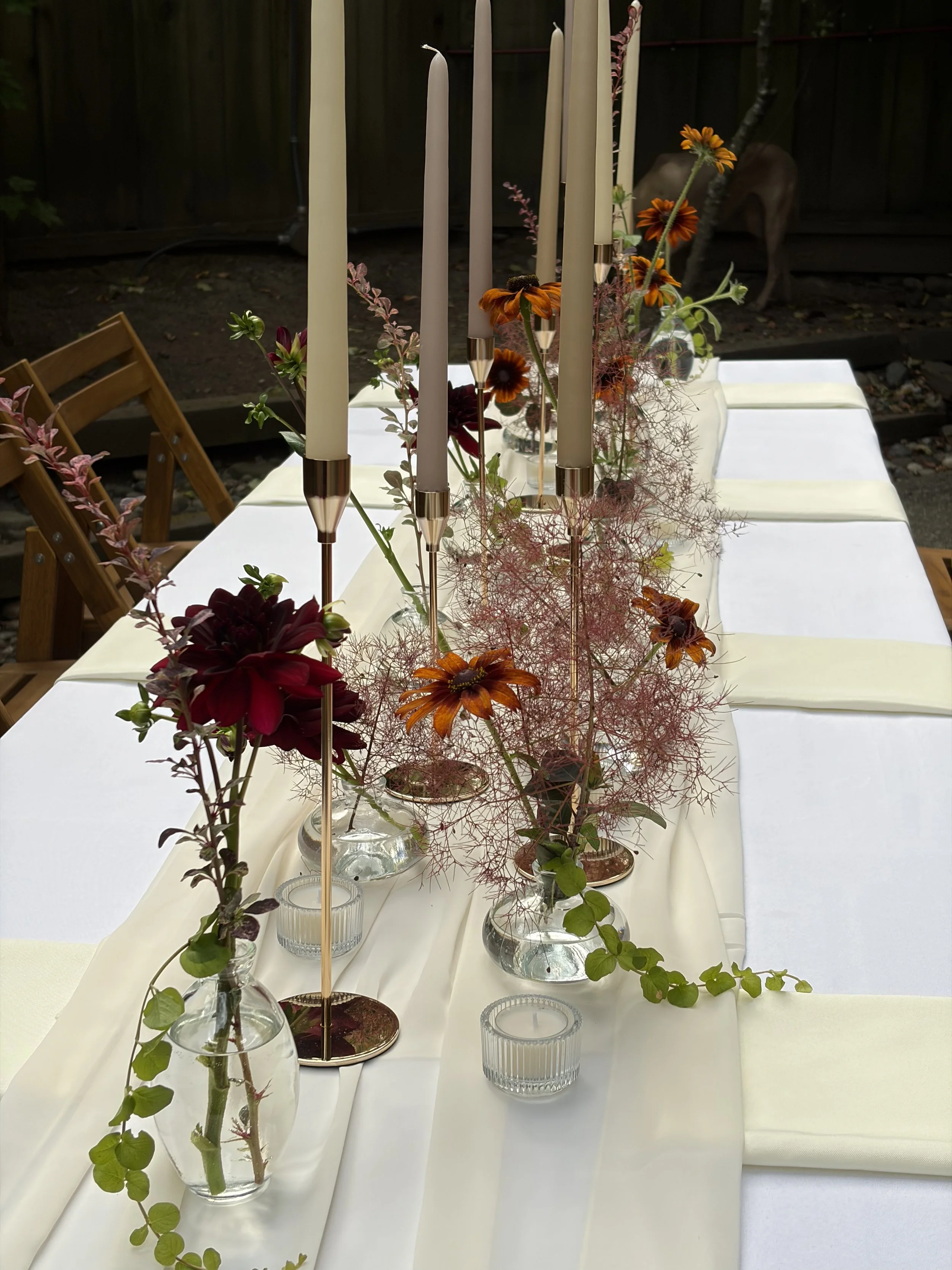 A table decorated with a white tablecloth, a floral centerpiece with orange, red, and pink flowers, surrounded by small glass vases with green foliage, tall white candles in gold and silver holders, and small glass candle holders, set against a backg