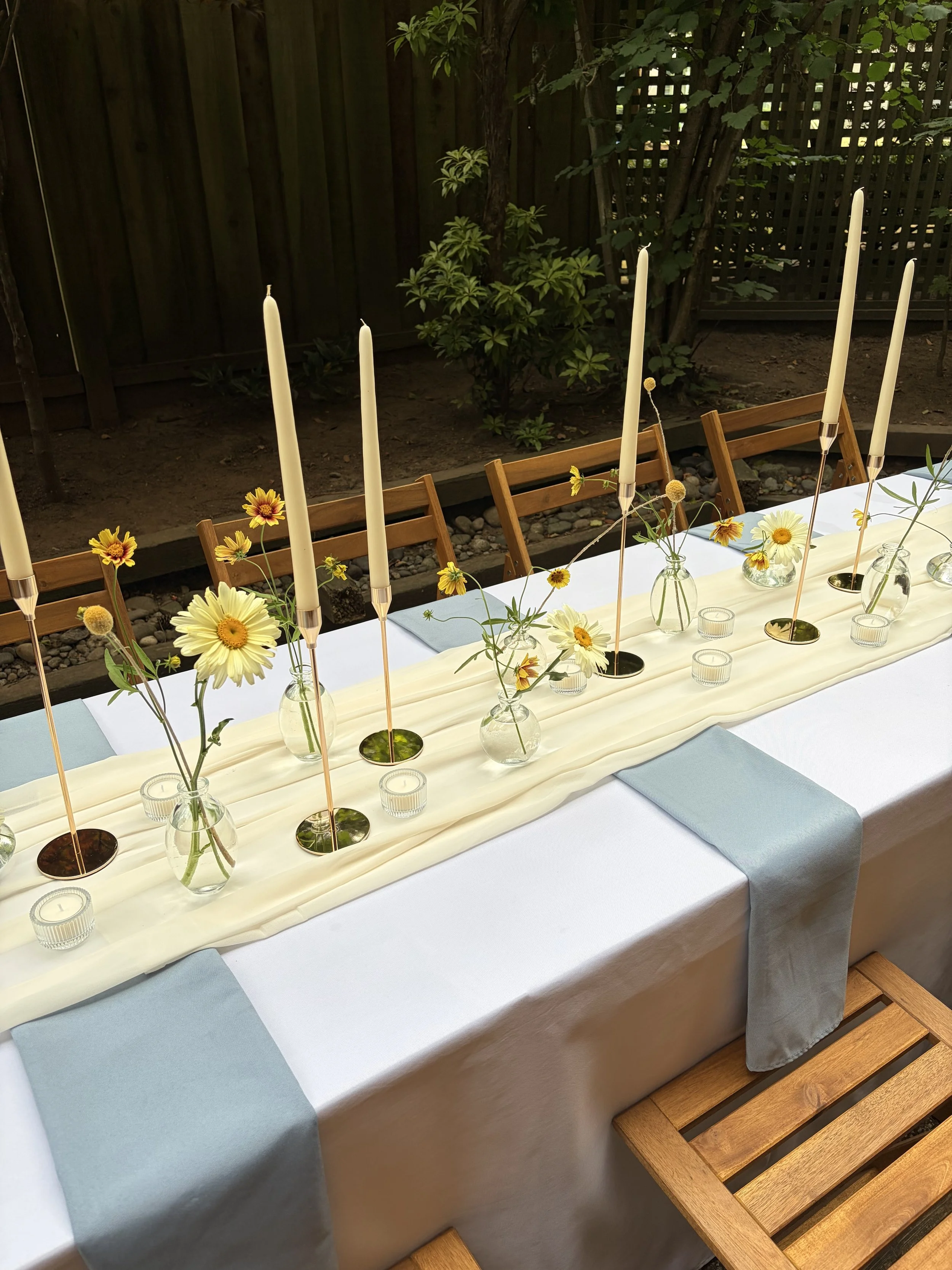 A decorated dining table with a white tablecloth, blue napkins, small glass vases with yellow and white flowers, tall white candles, and small candles in glass holders, set outdoors against a wooden fence and greenery.