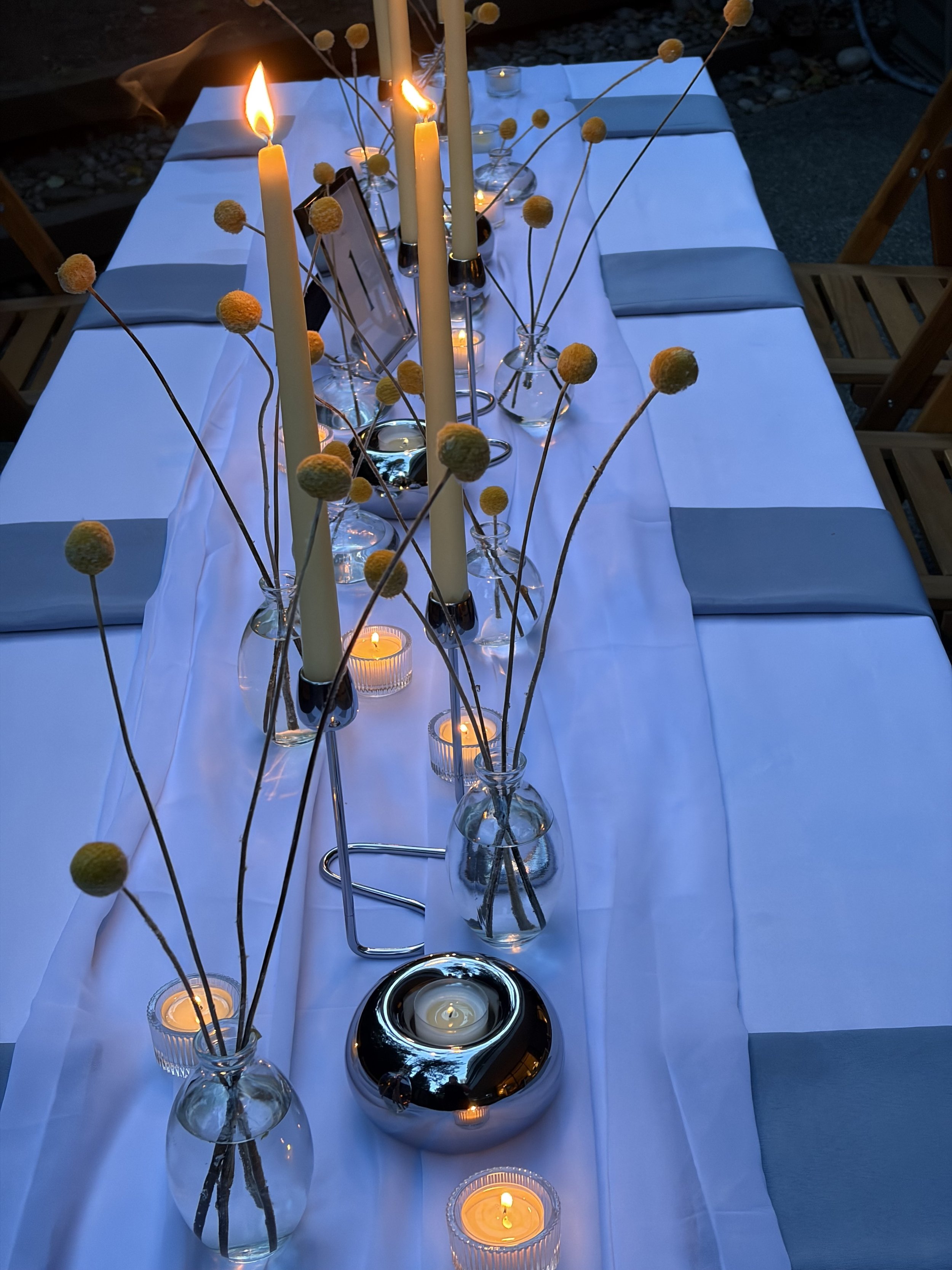 A long dining table set outdoors with a white tablecloth, decorated with lit candles in glass holders, small vases with yellow decorative pom-pom sticks, and candles in the background.