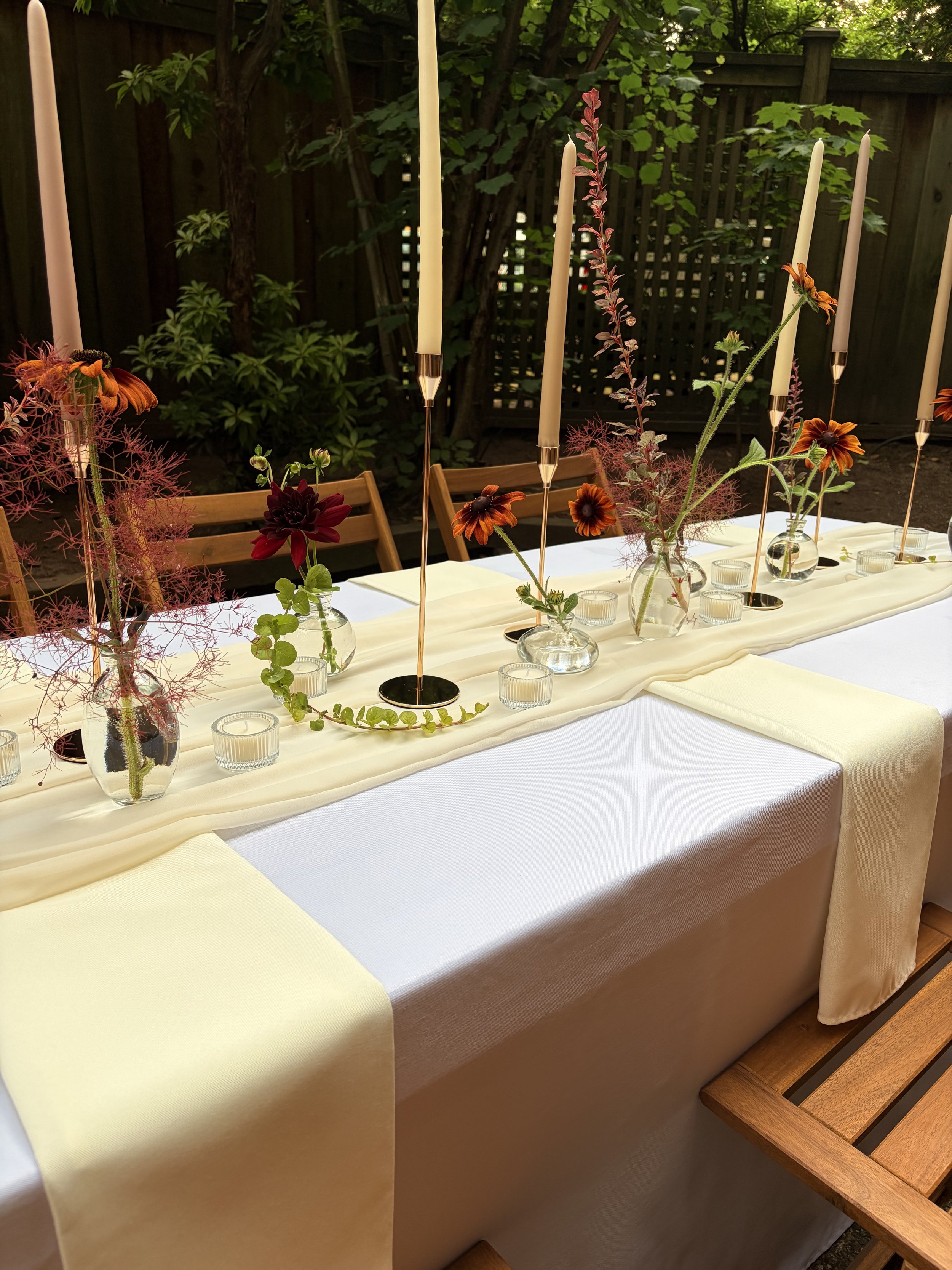 A long banquet table decorated with vases of flowers and tall white candles, set outdoors with a wooden privacy fence and green trees in the background.