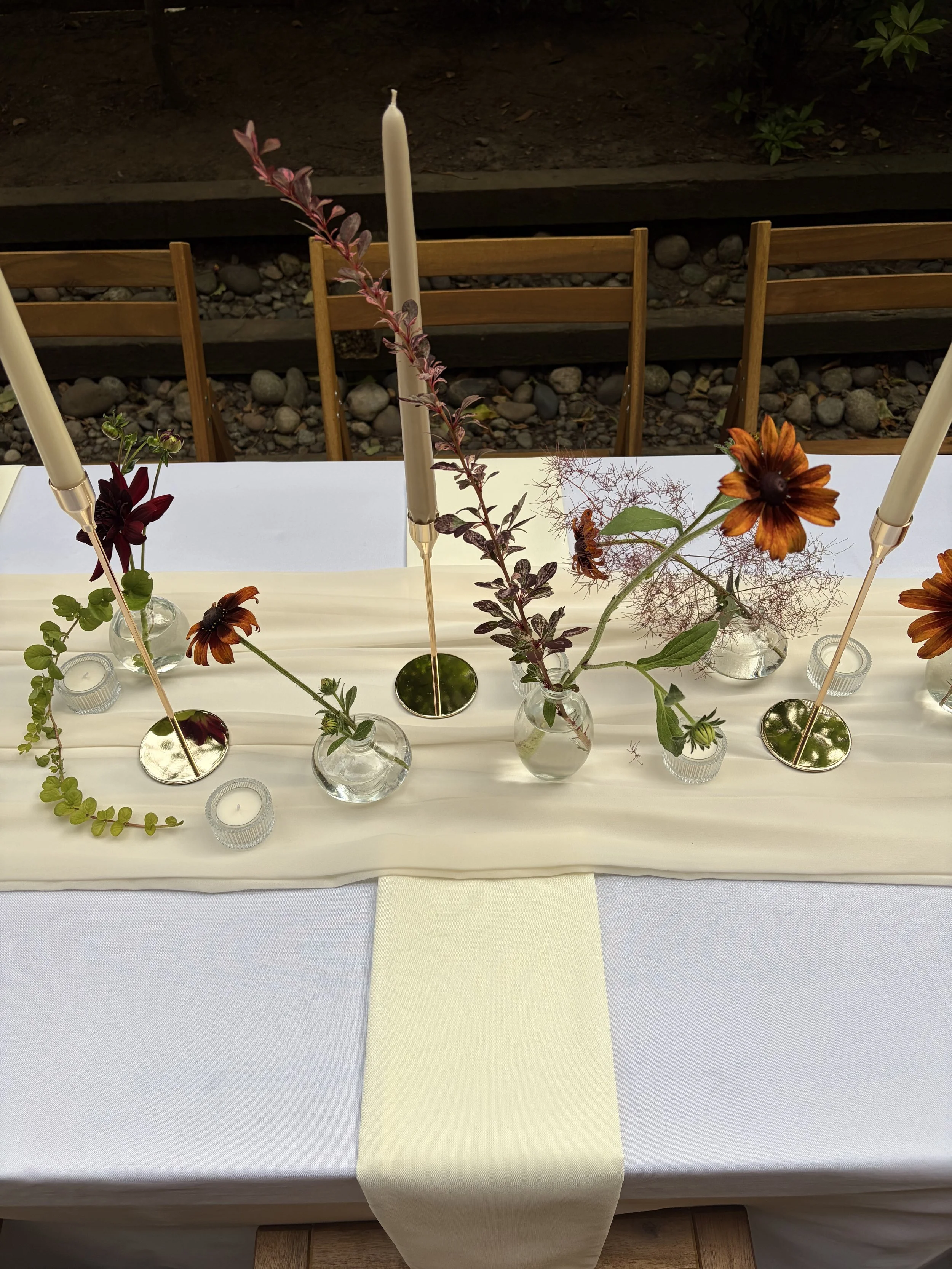 A table decorated with small vases of flowers, candles, and a cream-colored table runner, set outdoors with wooden chairs in the background.
