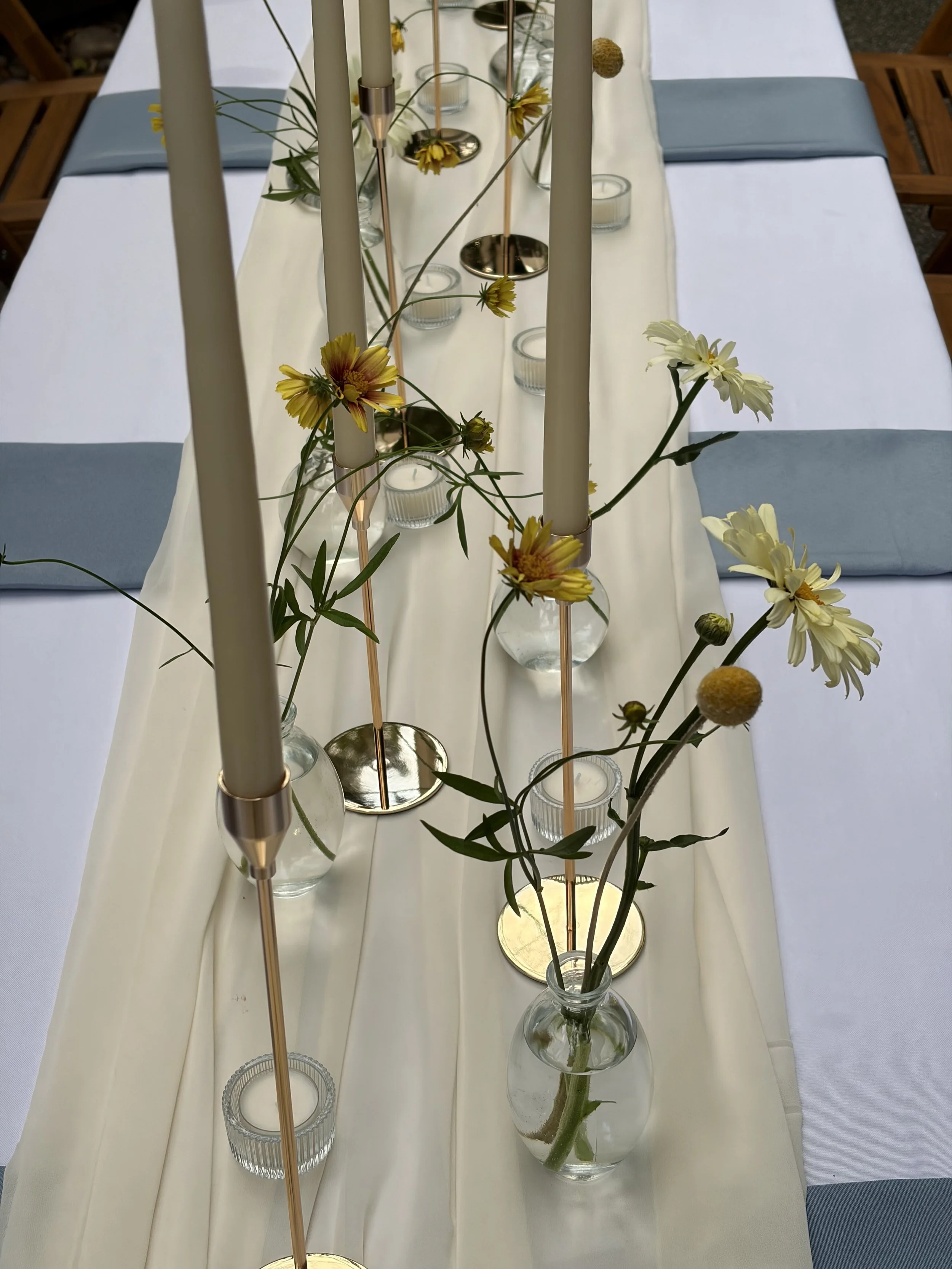 A table decorated with a white table runner, light blue napkins, glass candle holders, flowers in vases, and tall candles with candle holders.