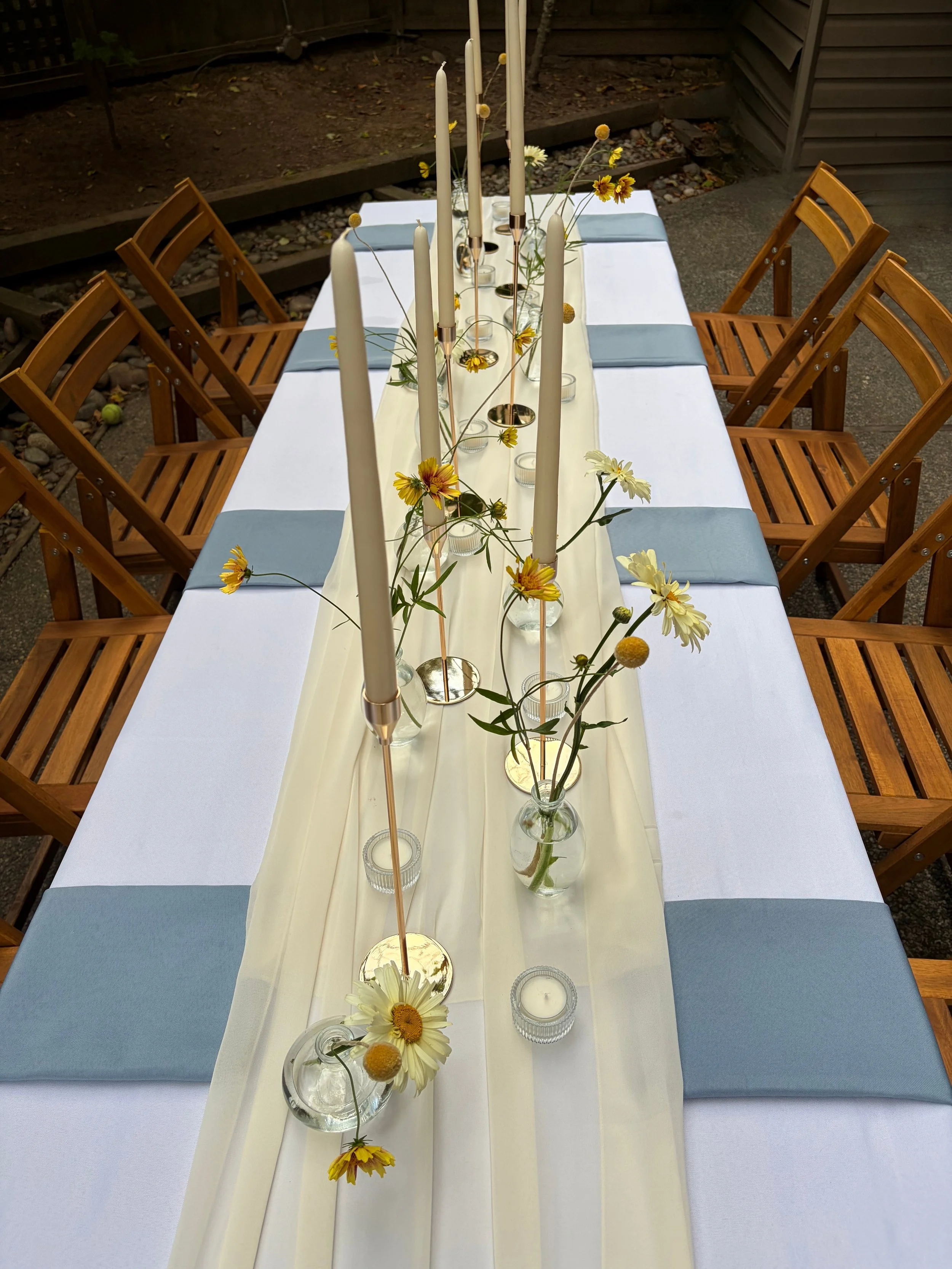 A long dining table decorated with candles and flowers, surrounded by wooden chairs, set outdoors on a patio.