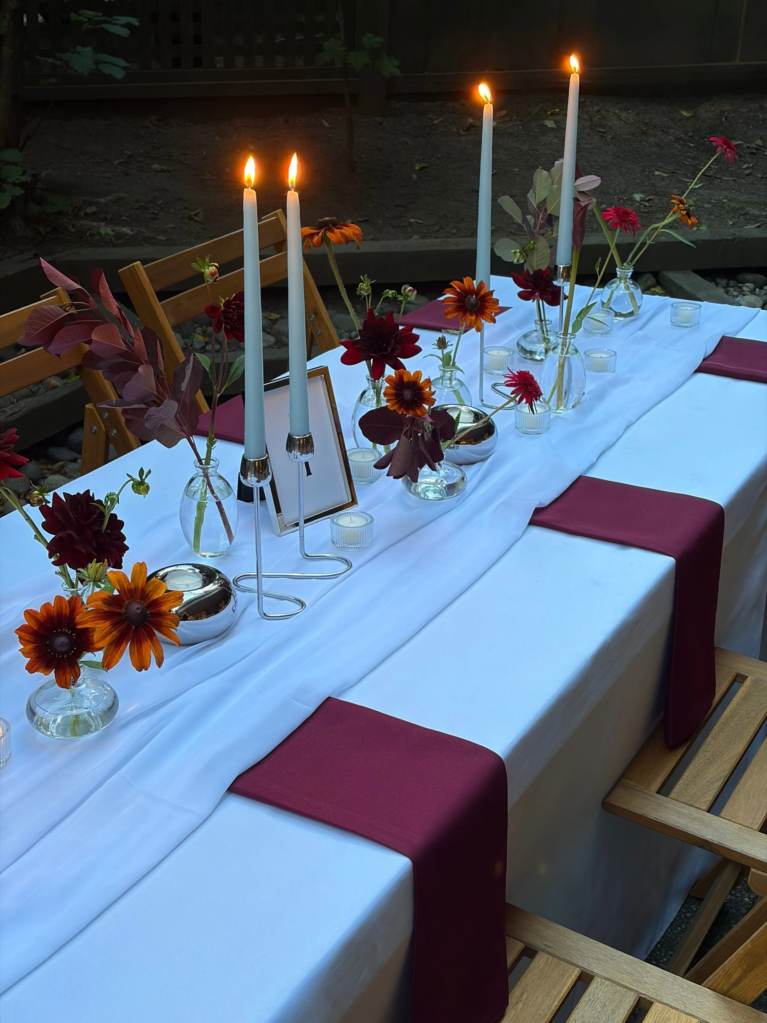 Decorated outdoor dining table with a white tablecloth, red napkins, vases of red and orange flowers, and tall white candles in candleholders, set for an evening meal.