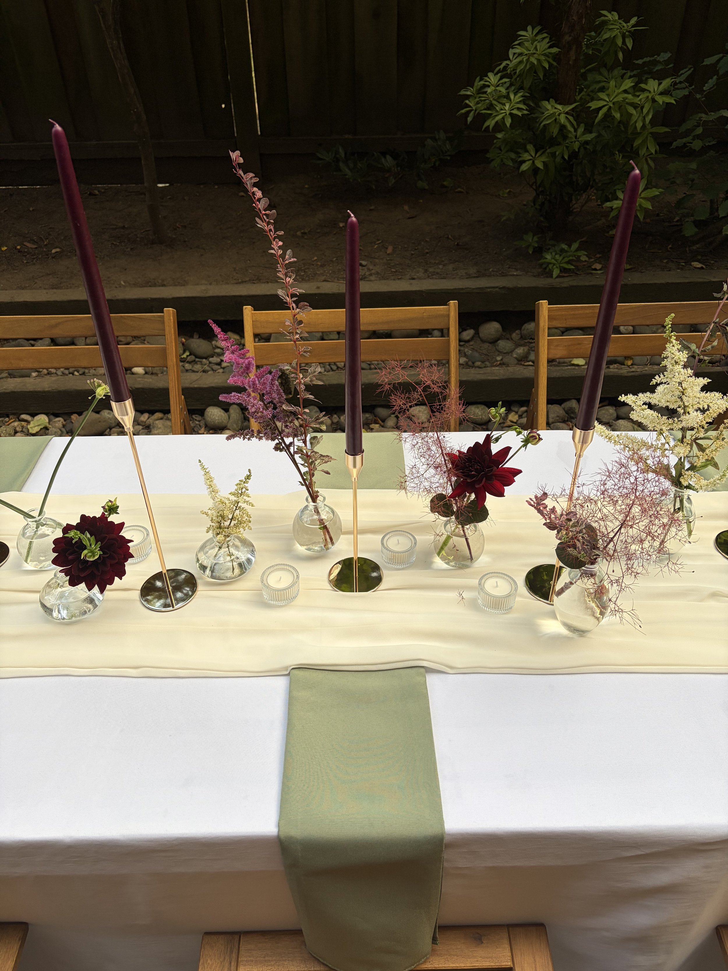 A dining table set with vases of flowers, purple candles, and small candles, with a light green table runner in a cozy outdoor setting.