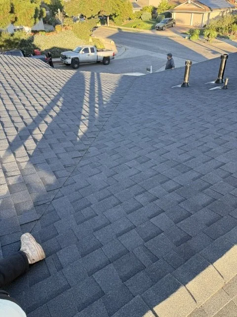 View of a residential roof with asphalt shingles, rapper and pipes, with a person sitting near the roof edge and a person walking on the street below.