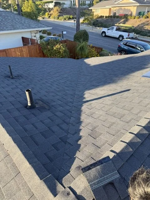 View of a house roof with two different shades of gray asphalt shingles, with a chimney pipe and ventilation pipe, overlooking a street with parked cars and a backyard with trees and shrubs.